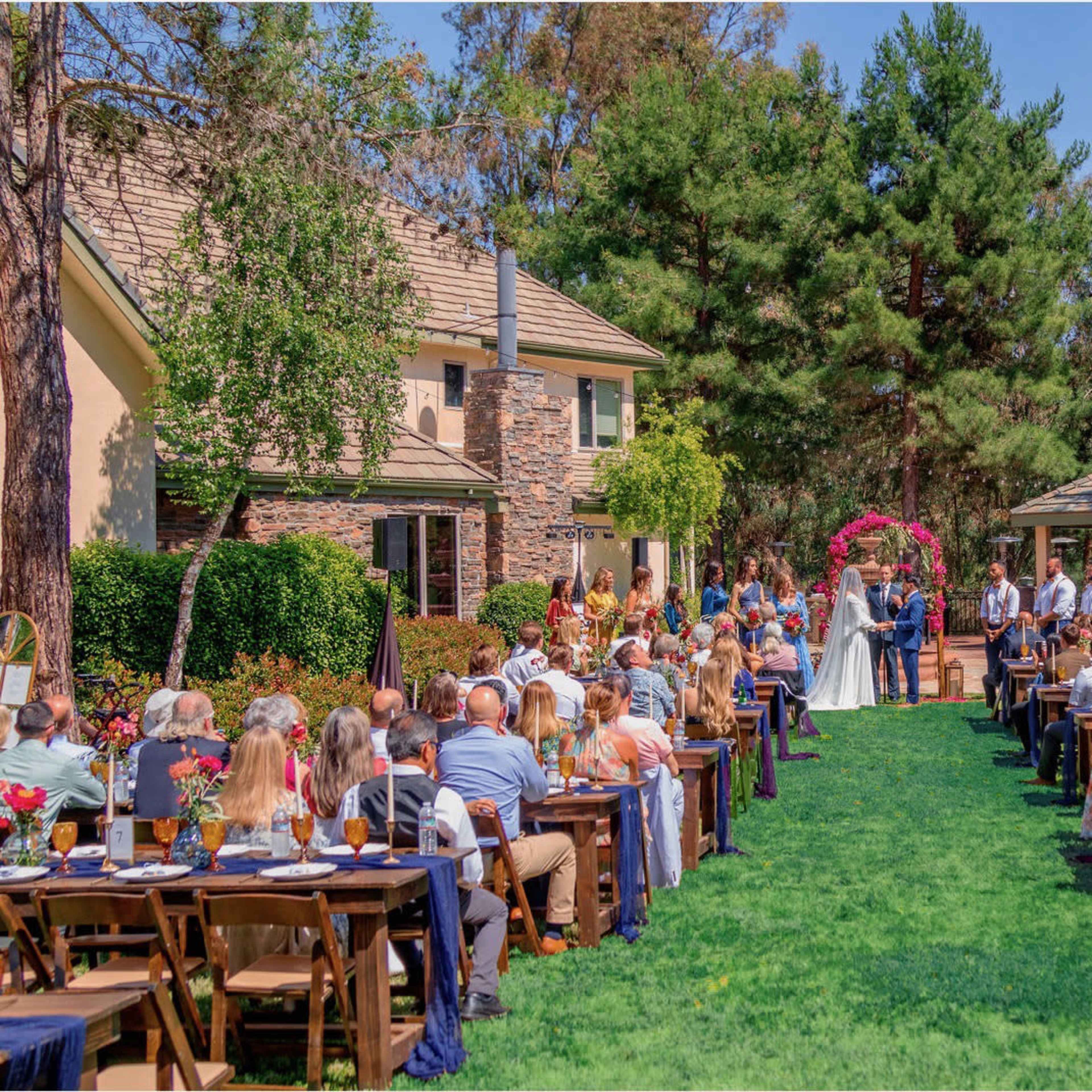 A wedding ceremony takes place outdoors in a garden setting, with guests seated on either side of an aisle leading to the couple at an altar adorned with flowers.