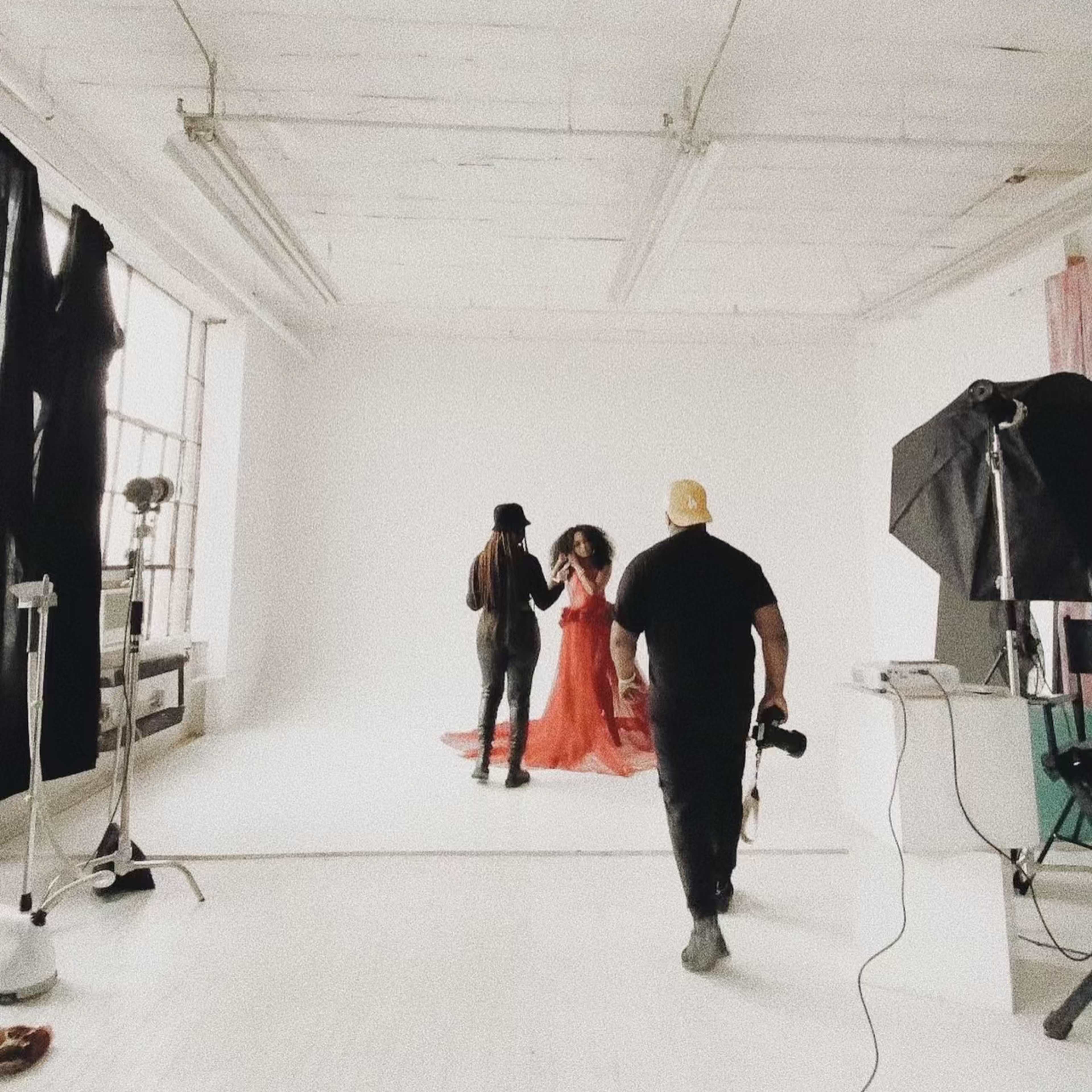 A photoshoot in a spacious studio, where a model in a red dress is being attended to by two individuals, while a camera is positioned nearby.