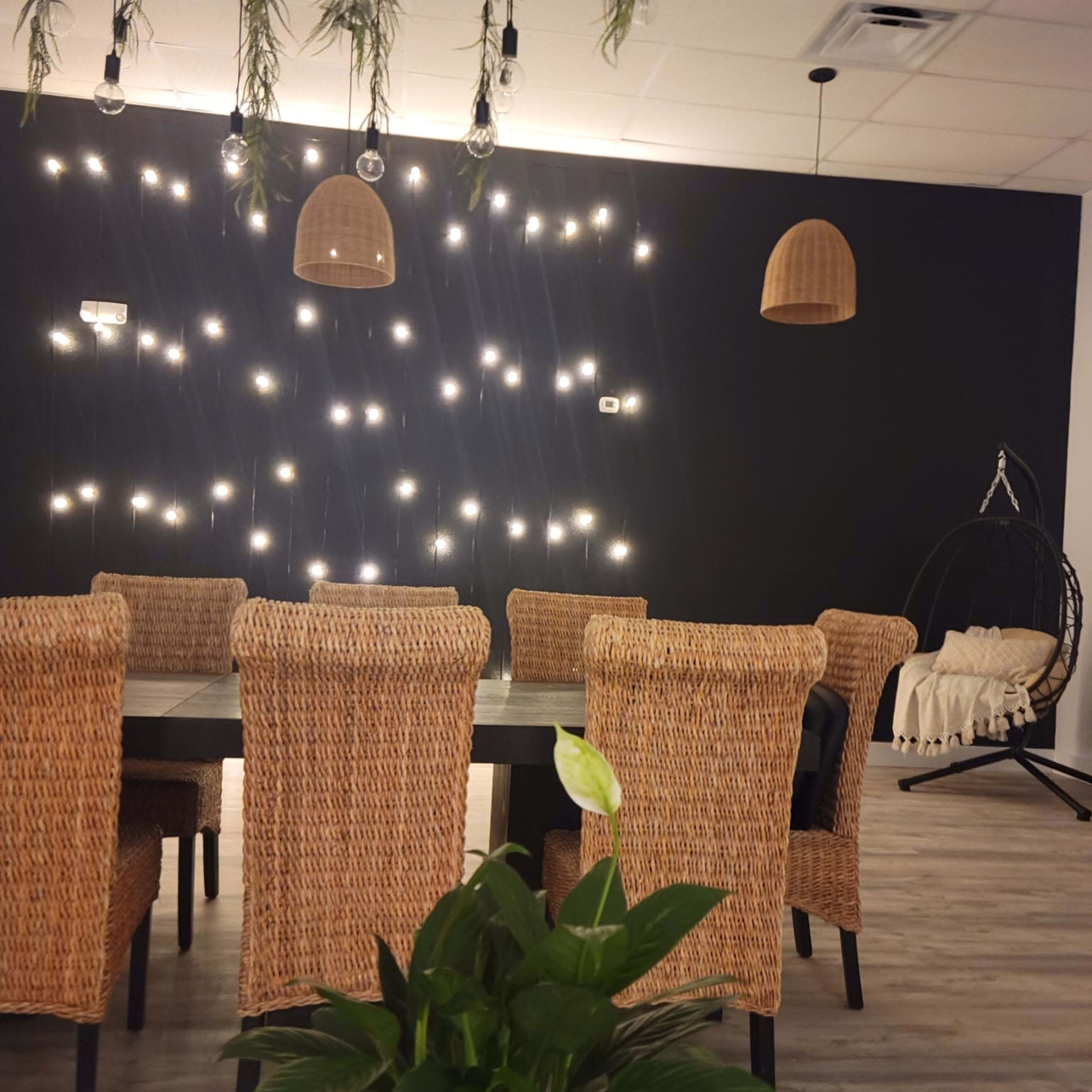 A dining area features wicker chairs around a dark table, with hanging lights and a plant in the foreground.