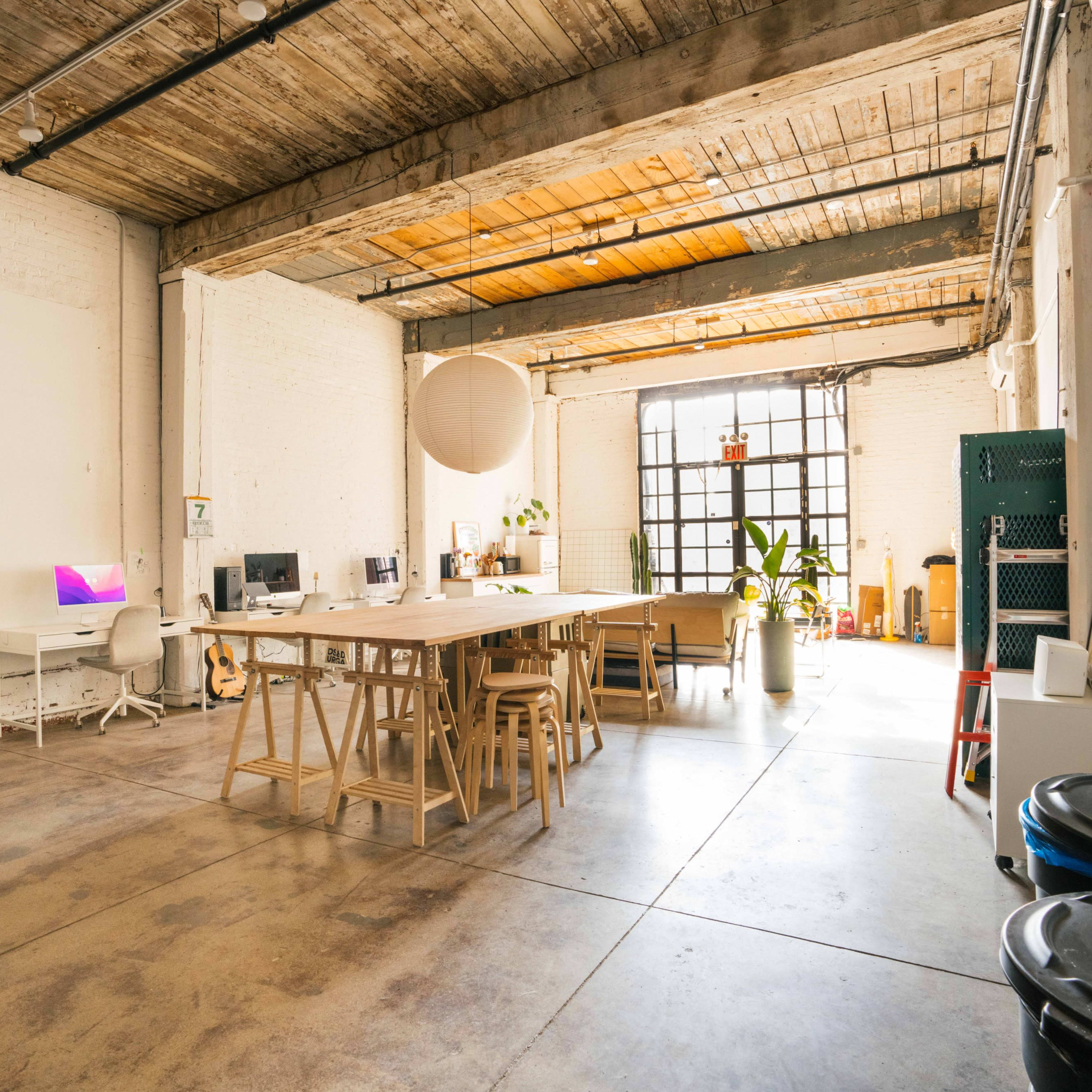 A spacious, well-lit office area with a long wooden table, several workstations, and large windows allowing natural light in.