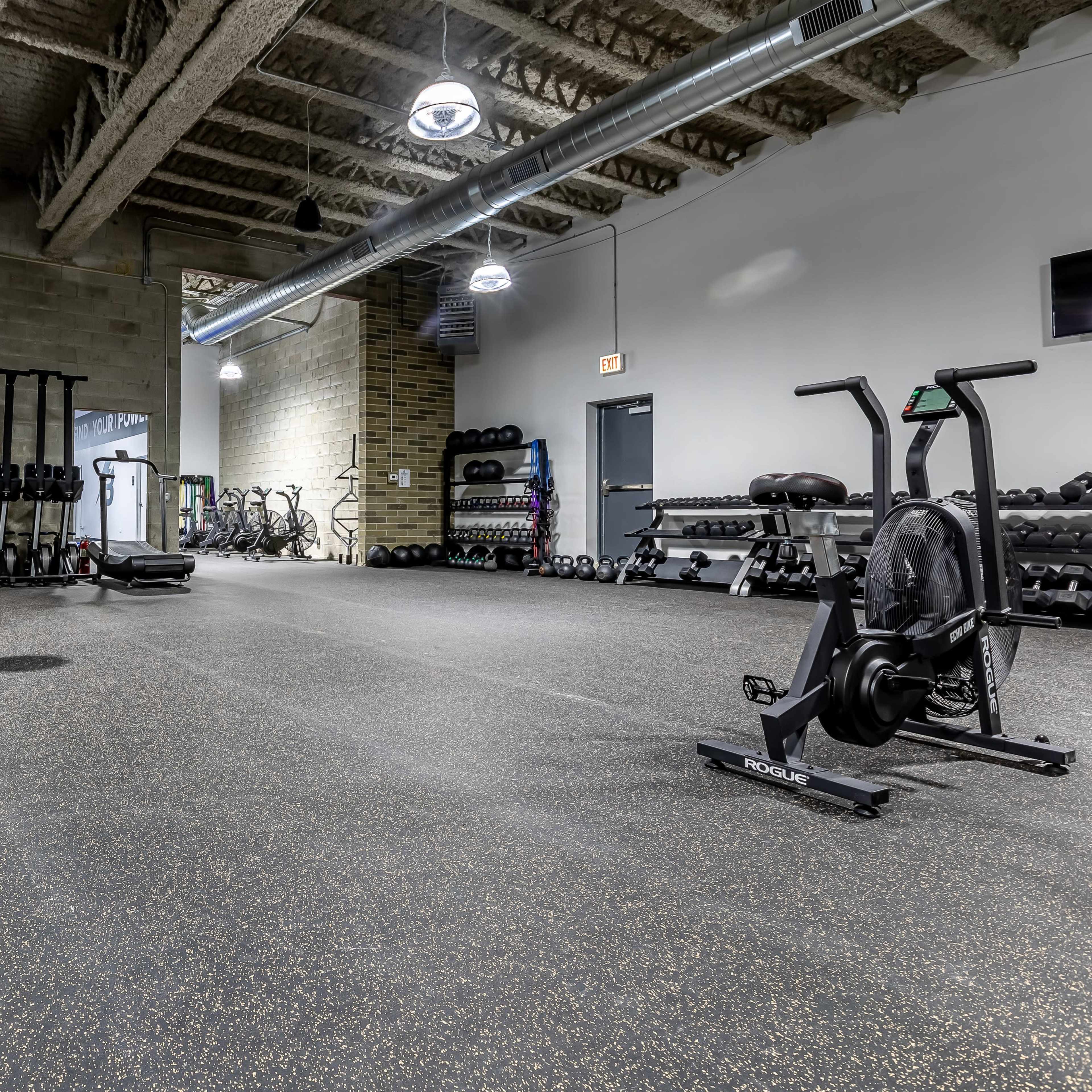 A spacious gym interior with various workout equipment, including weights, a stationary bike, and a sled, on a rubberized floor.
