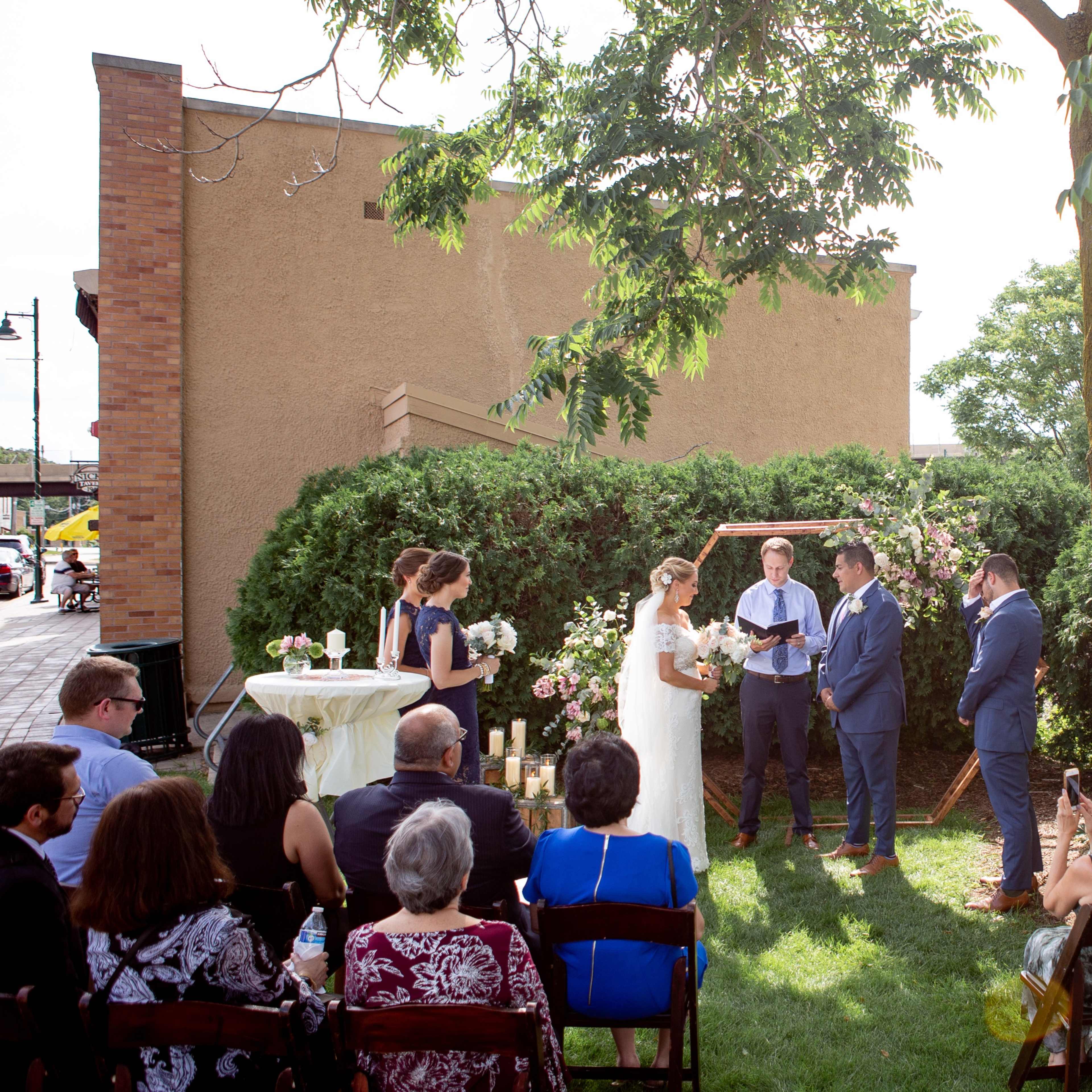 A couple stands under a wooden arch adorned with flowers while exchanging vows in a garden setting, surrounded by guests seated on chairs.