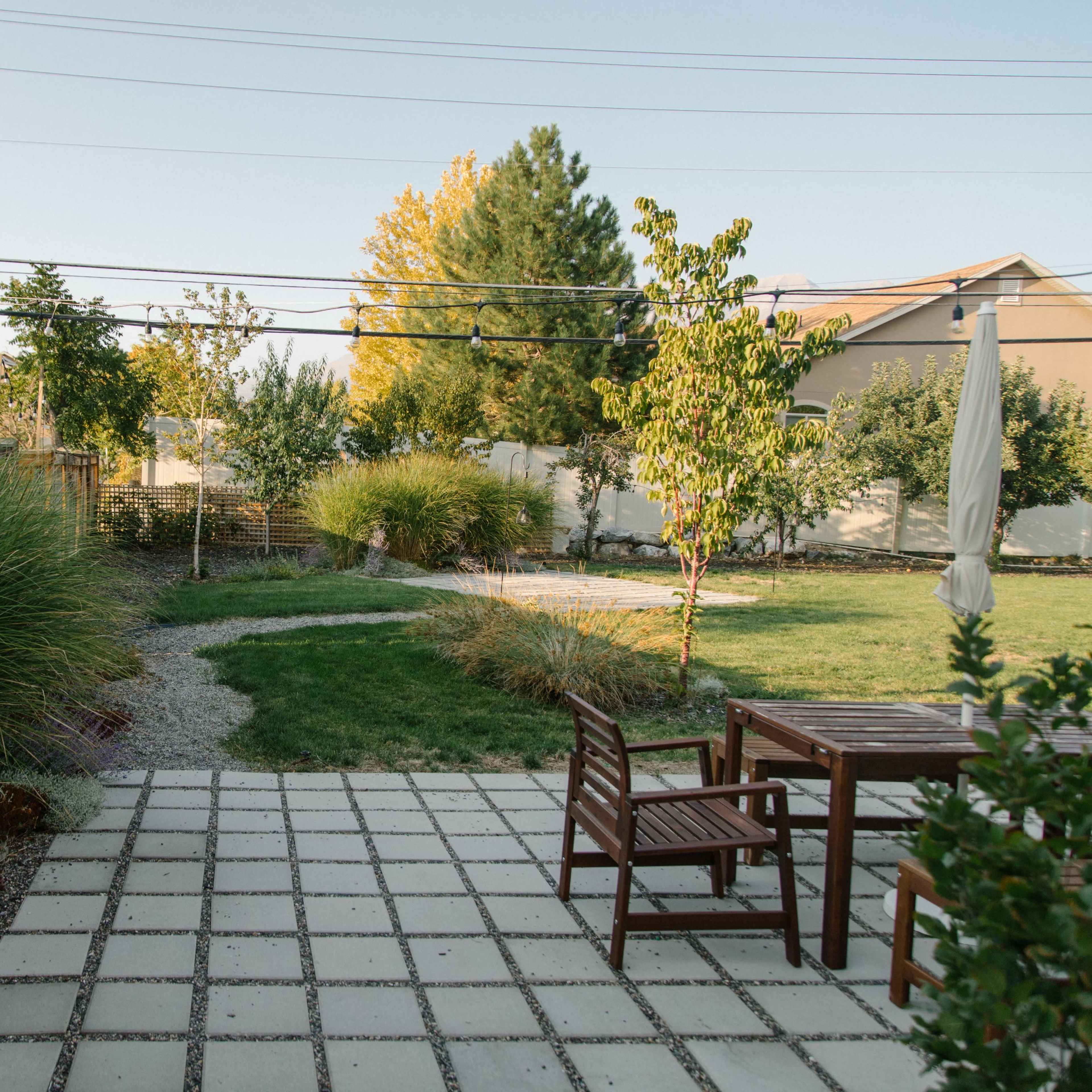 A backyard landscape features a stone patio with wooden furniture, surrounded by gravel paths, ornamental grasses, and trees.