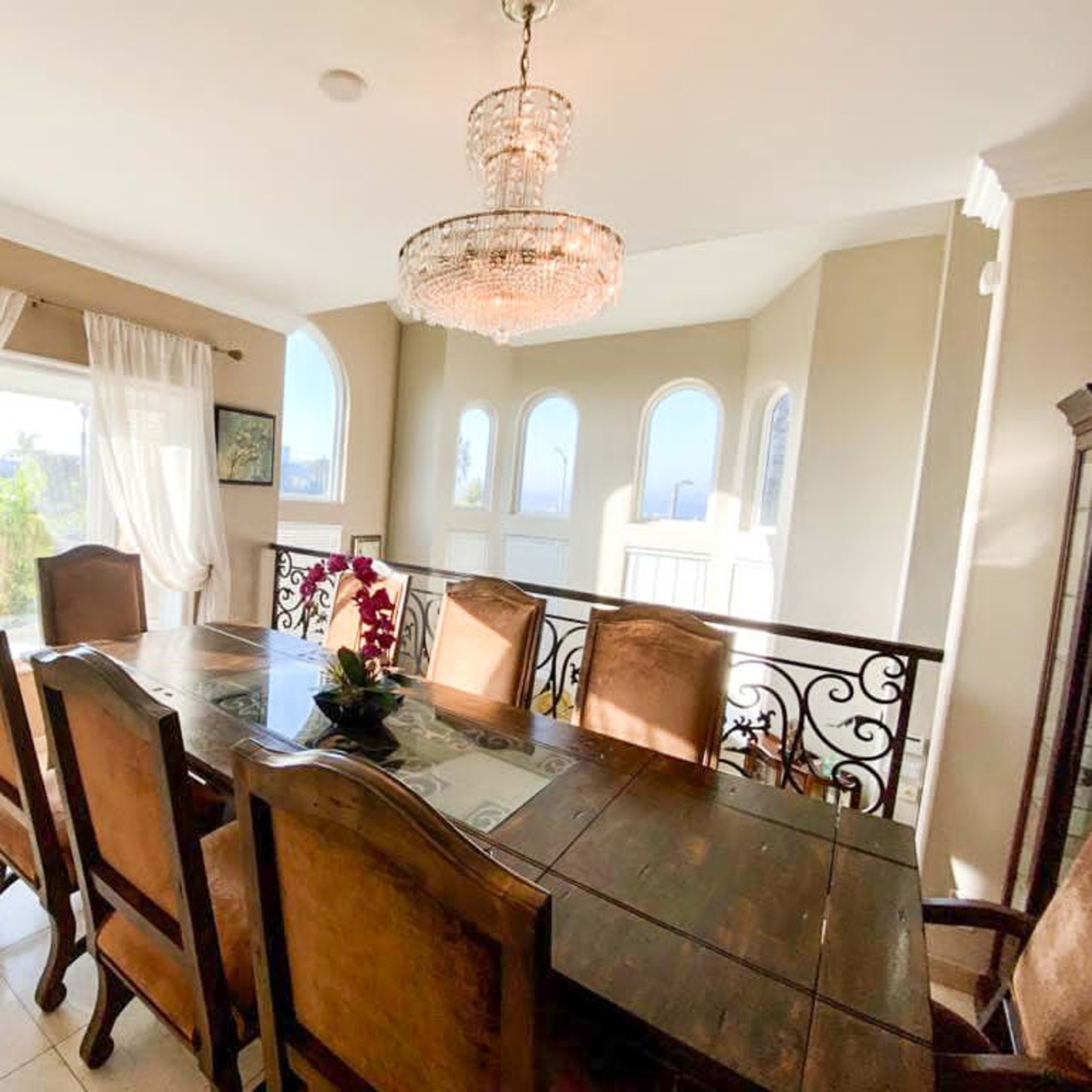 A wooden dining table surrounded by eight chairs is set in a room with large windows and a chandelier overhead.