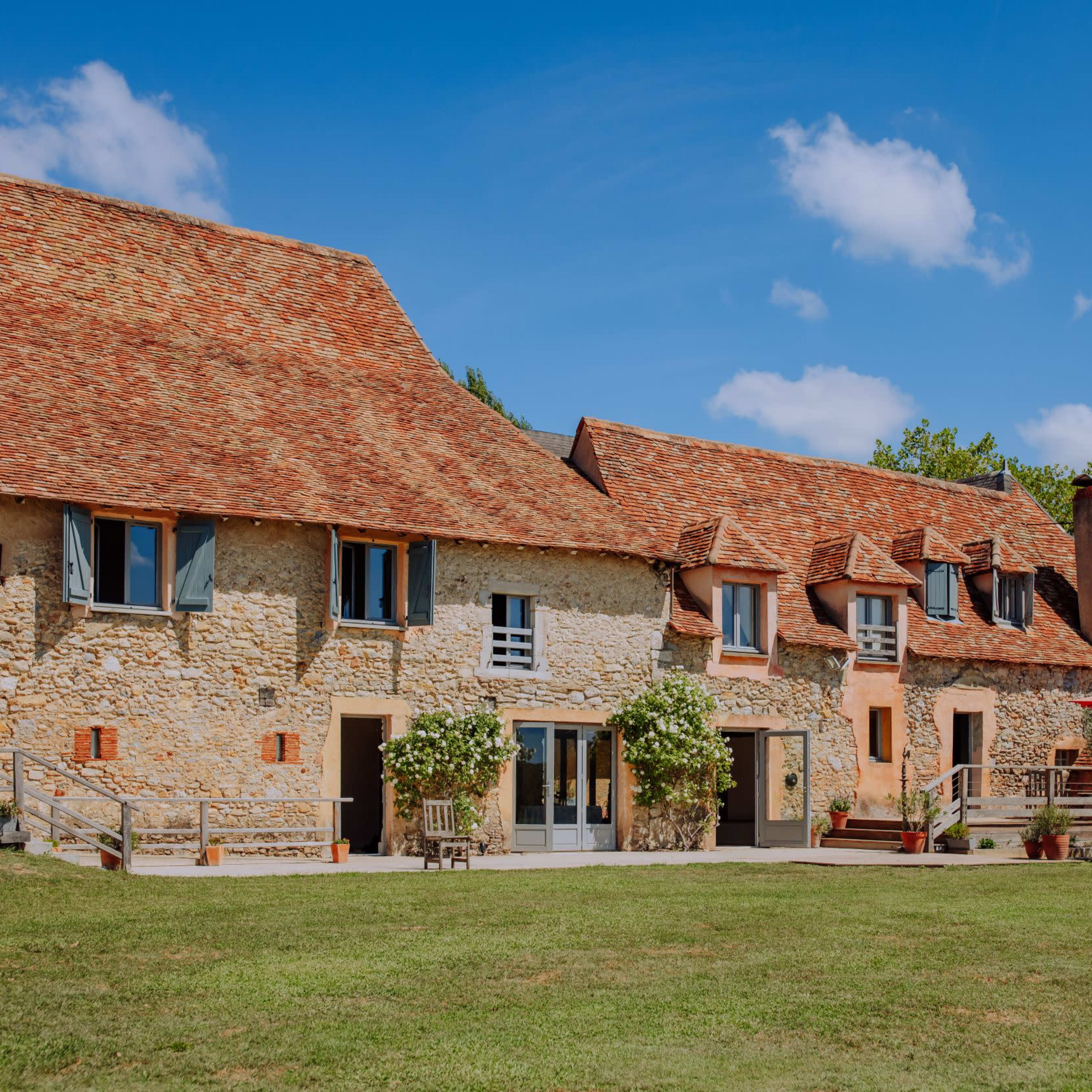 A stone house with a red-tiled roof and a grassy lawn is set against a blue sky with scattered clouds.
