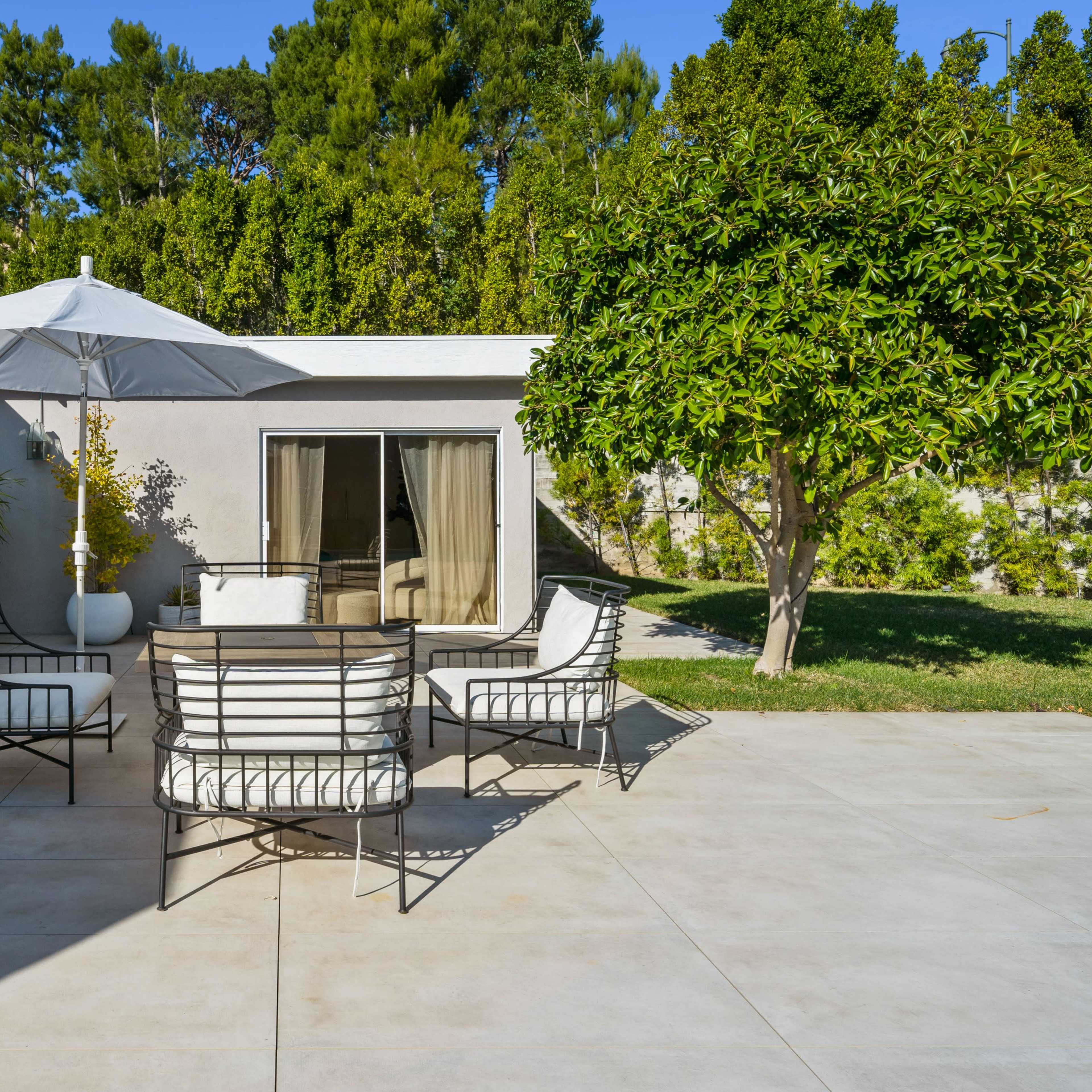 A patio area features a table and four chairs beneath a large umbrella, surrounded by greenery and sliding glass doors to the house.