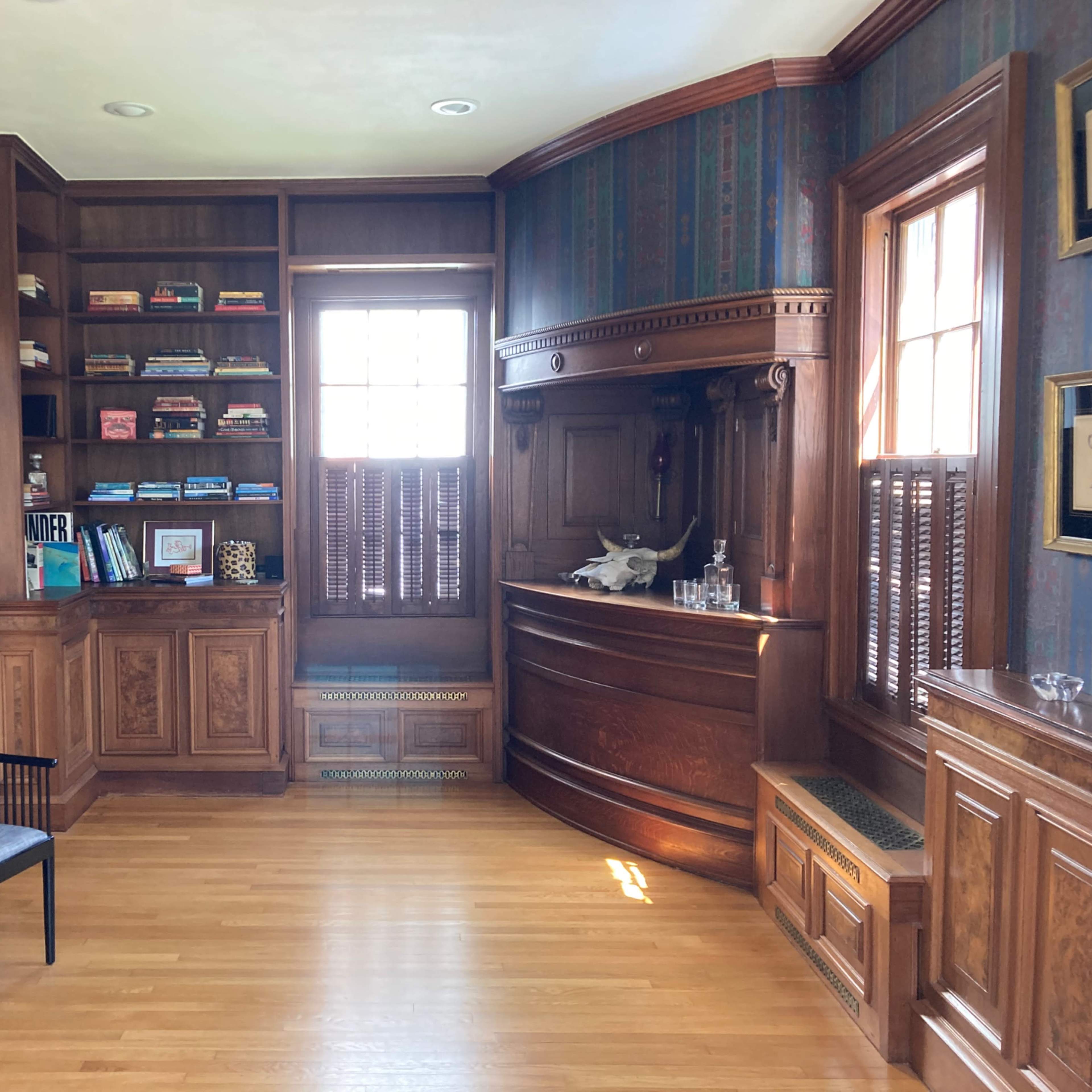 The room features wooden paneling, a built-in bookshelf filled with books, a bar area, and a window with wooden shutters.