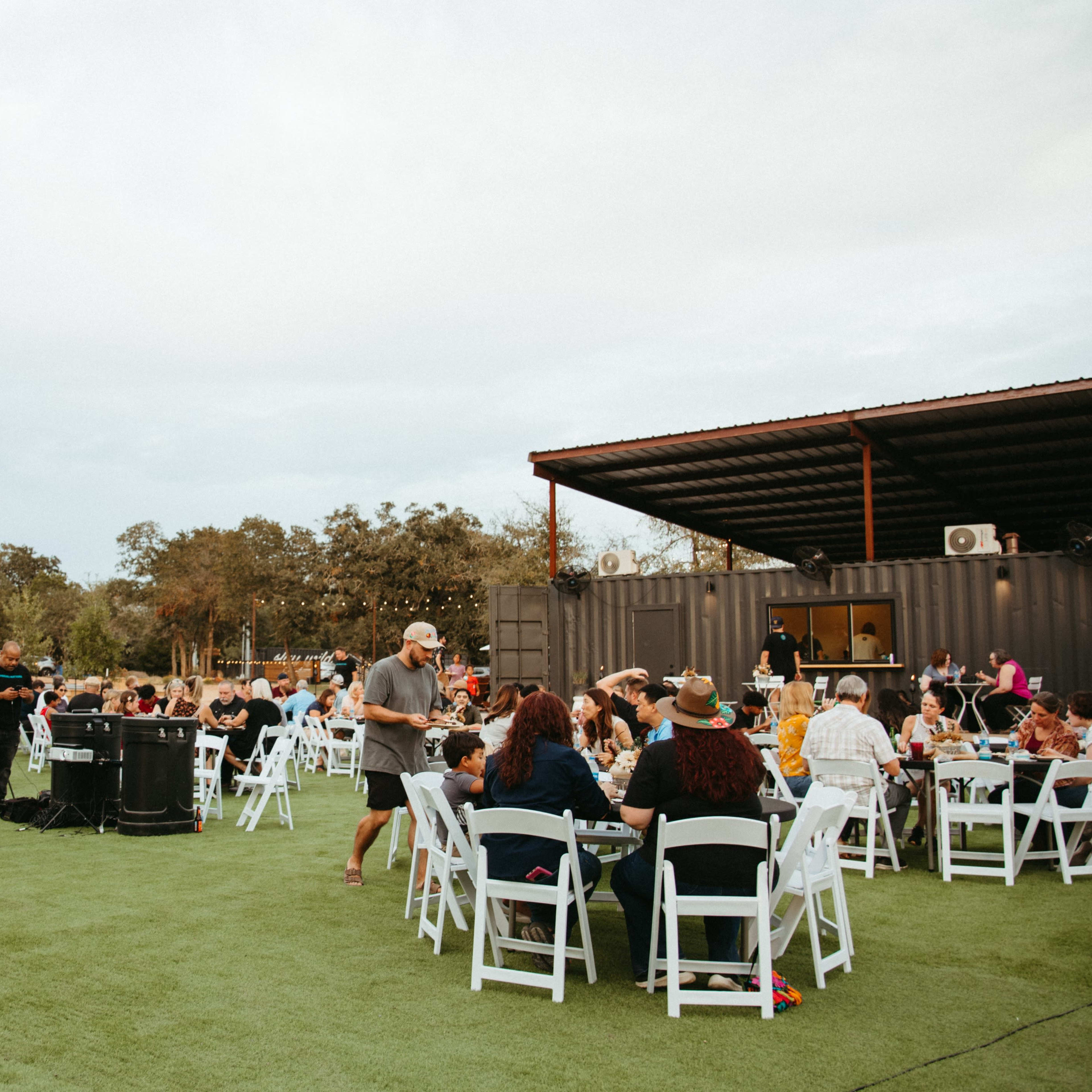 A large outdoor gathering features numerous tables filled with people, set up on green turf beneath a cloudy sky.