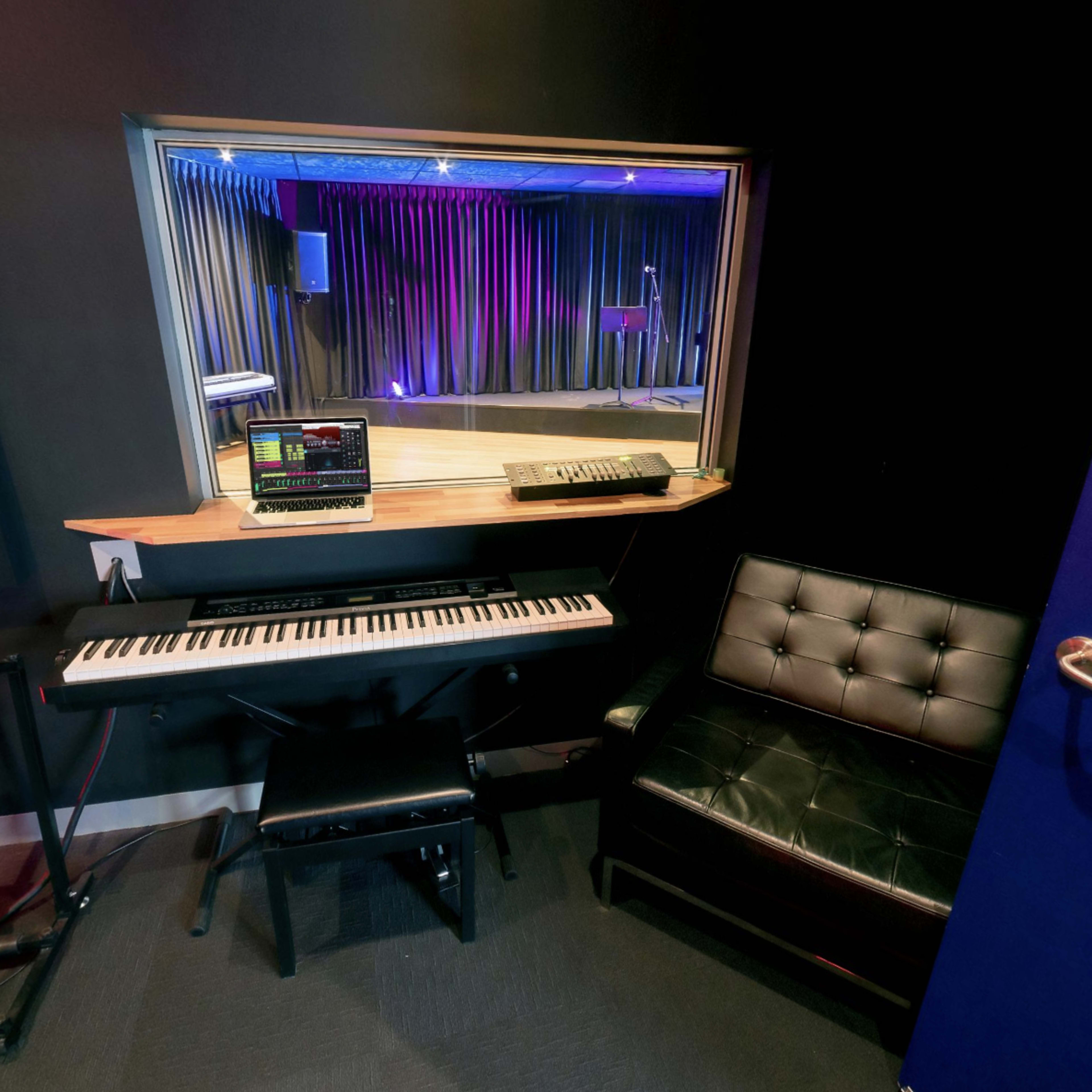 A small music studio corner featuring a keyboard, a computer on a desktop, a black leather chair, and acoustic guitars leaning against the wall.