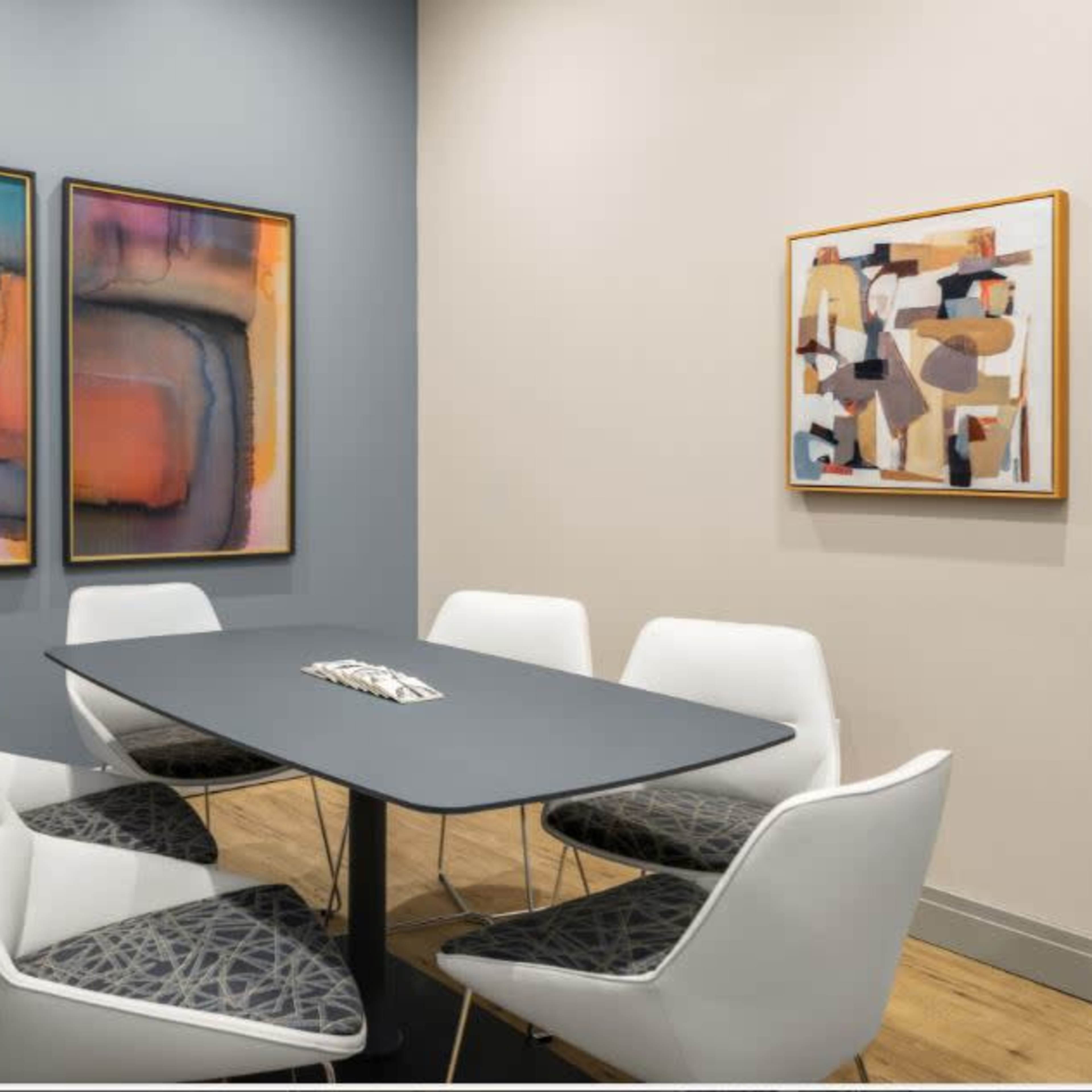 A modern meeting room with a gray table surrounded by six white chairs, featuring contemporary abstract paintings on the walls.