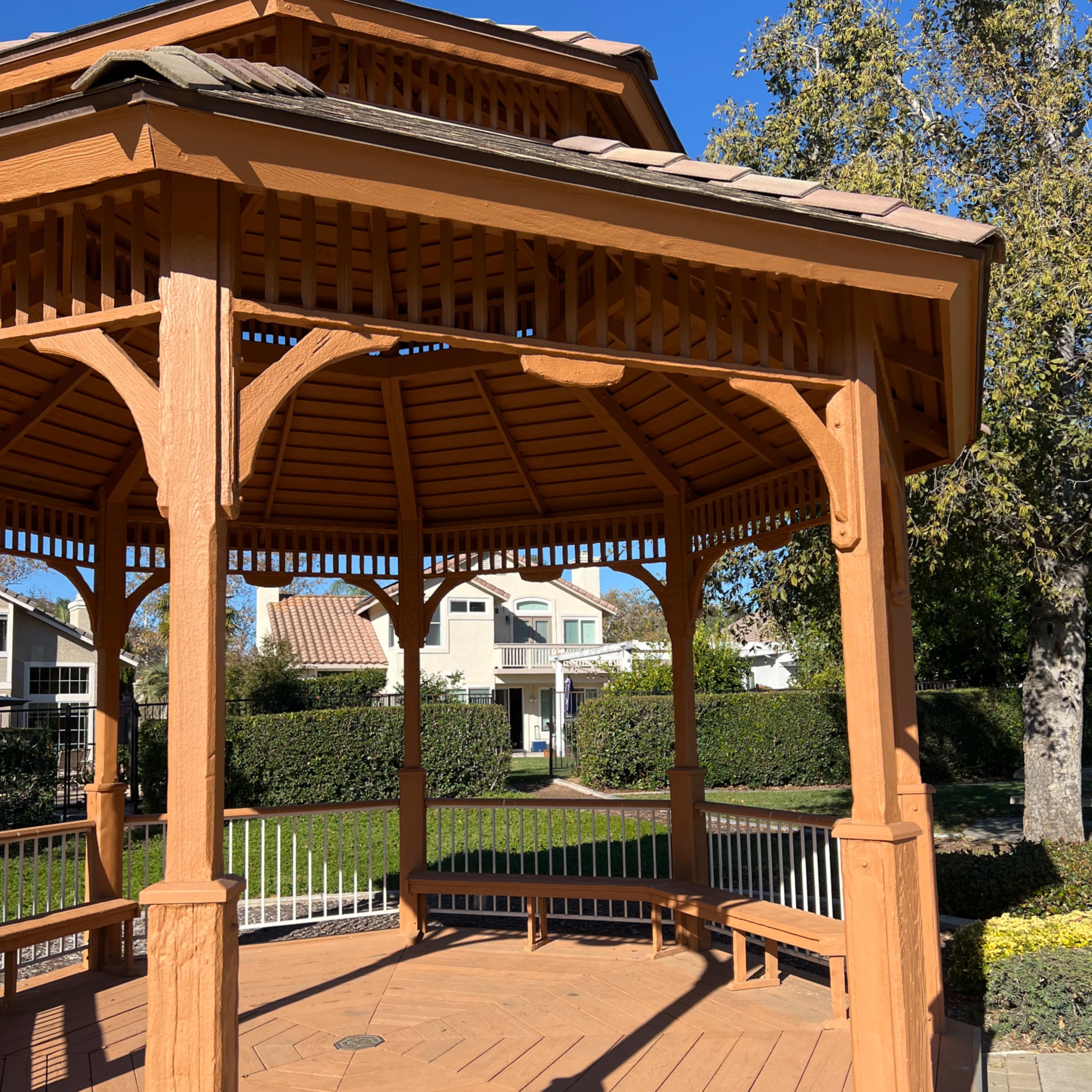 A wooden gazebo with a shingled roof stands in a park, surrounded by neatly trimmed hedges and nearby houses.