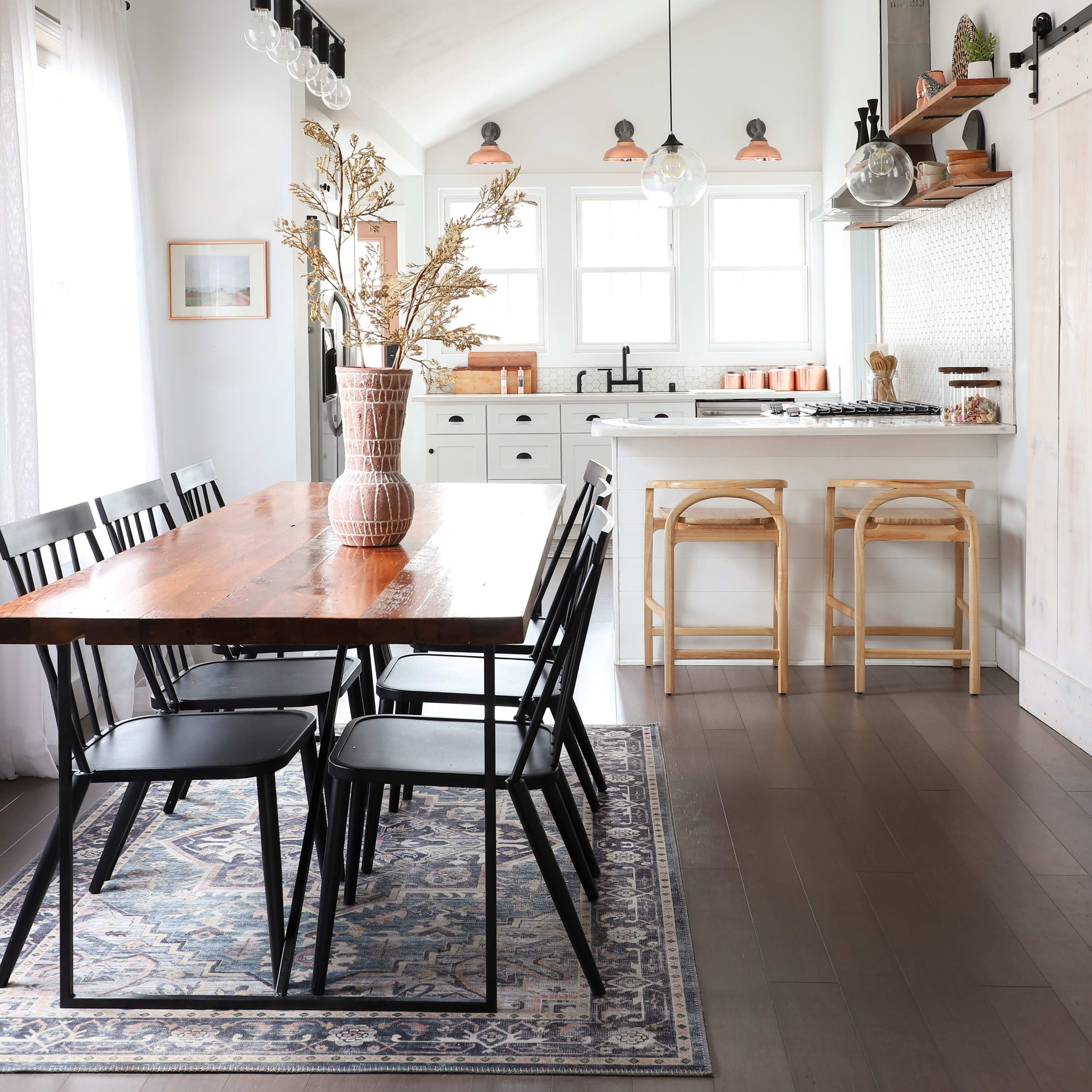 A modern kitchen features a wooden dining table surrounded by black chairs, with a bright decor including a vase of dried flowers and barstools at a counter.