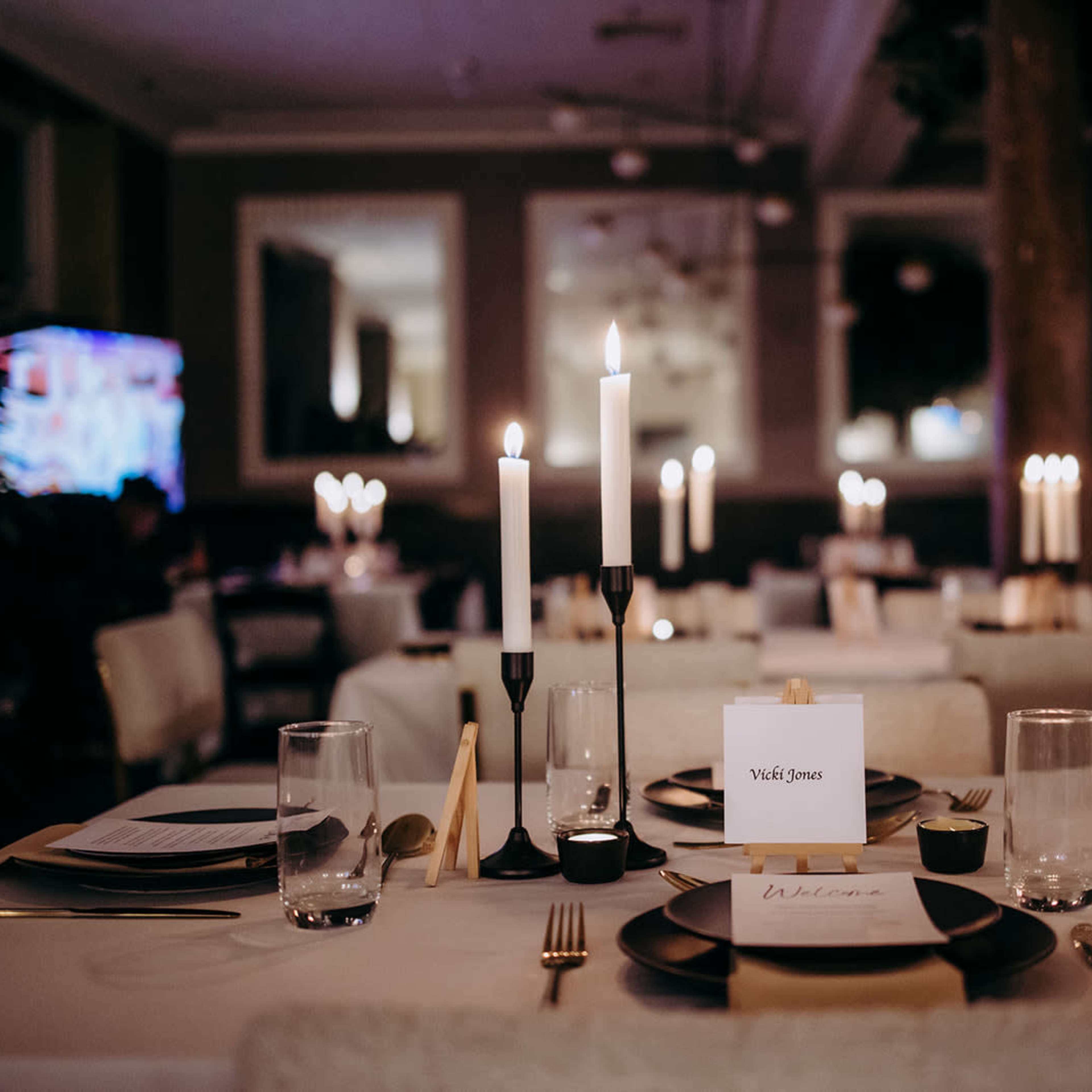 A neatly arranged dining area with tables set for an event, featuring candles, place cards, and glassware.
