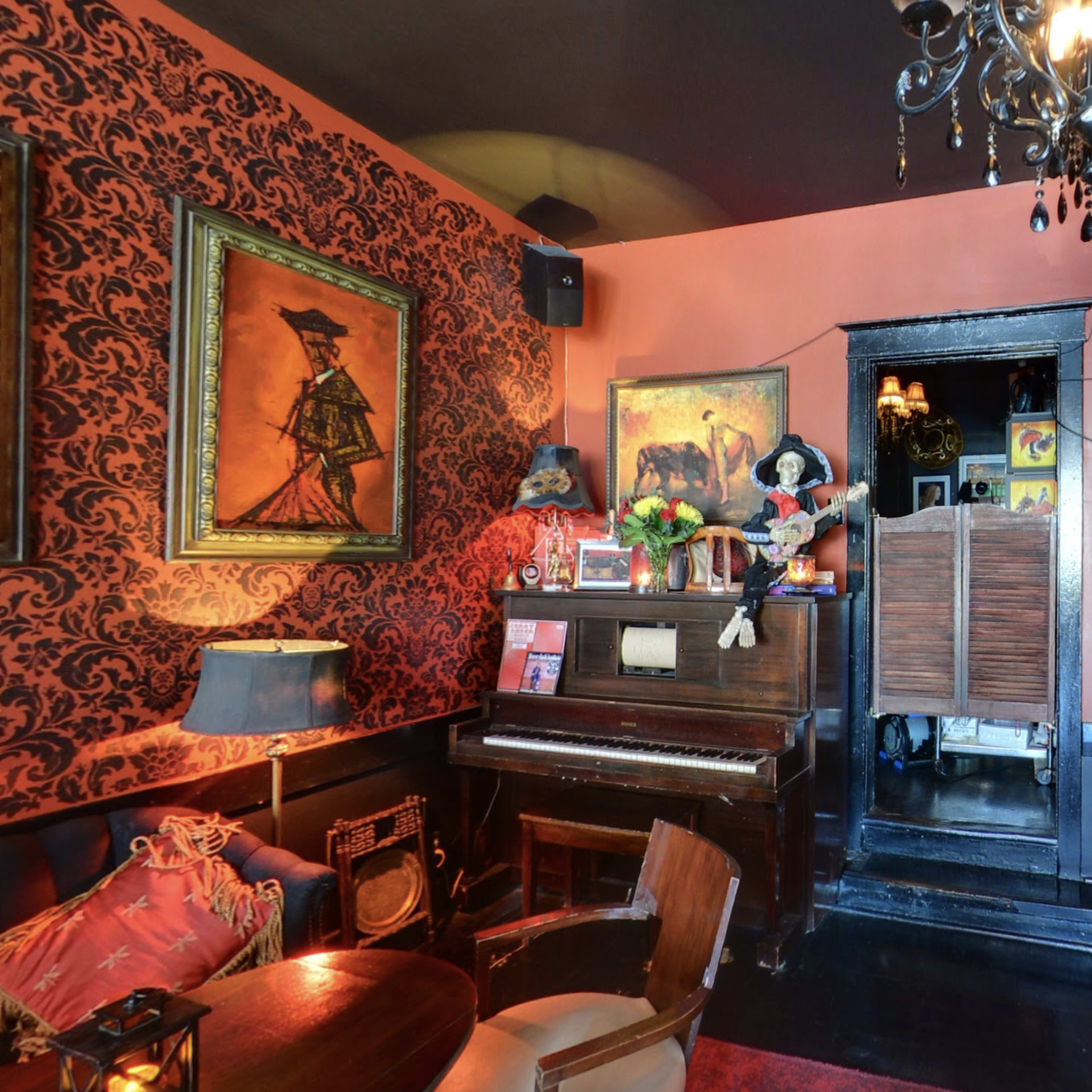 The interior of a cozy bar features dark walls adorned with framed artwork, a vintage piano, and a chandelier, creating an intimate atmosphere.