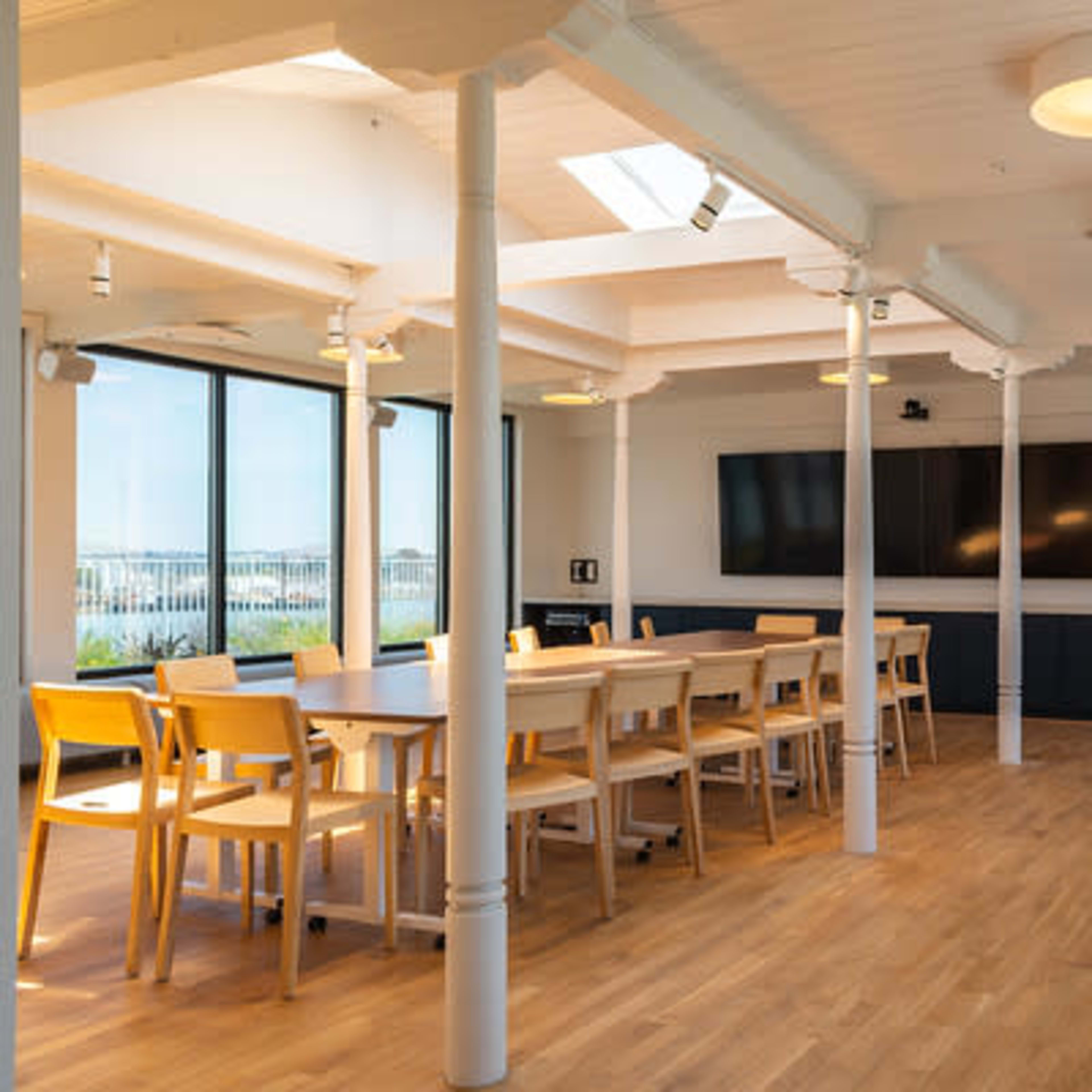 A spacious, modern meeting room with a long table surrounded by chairs, large windows, and a wall-mounted television.