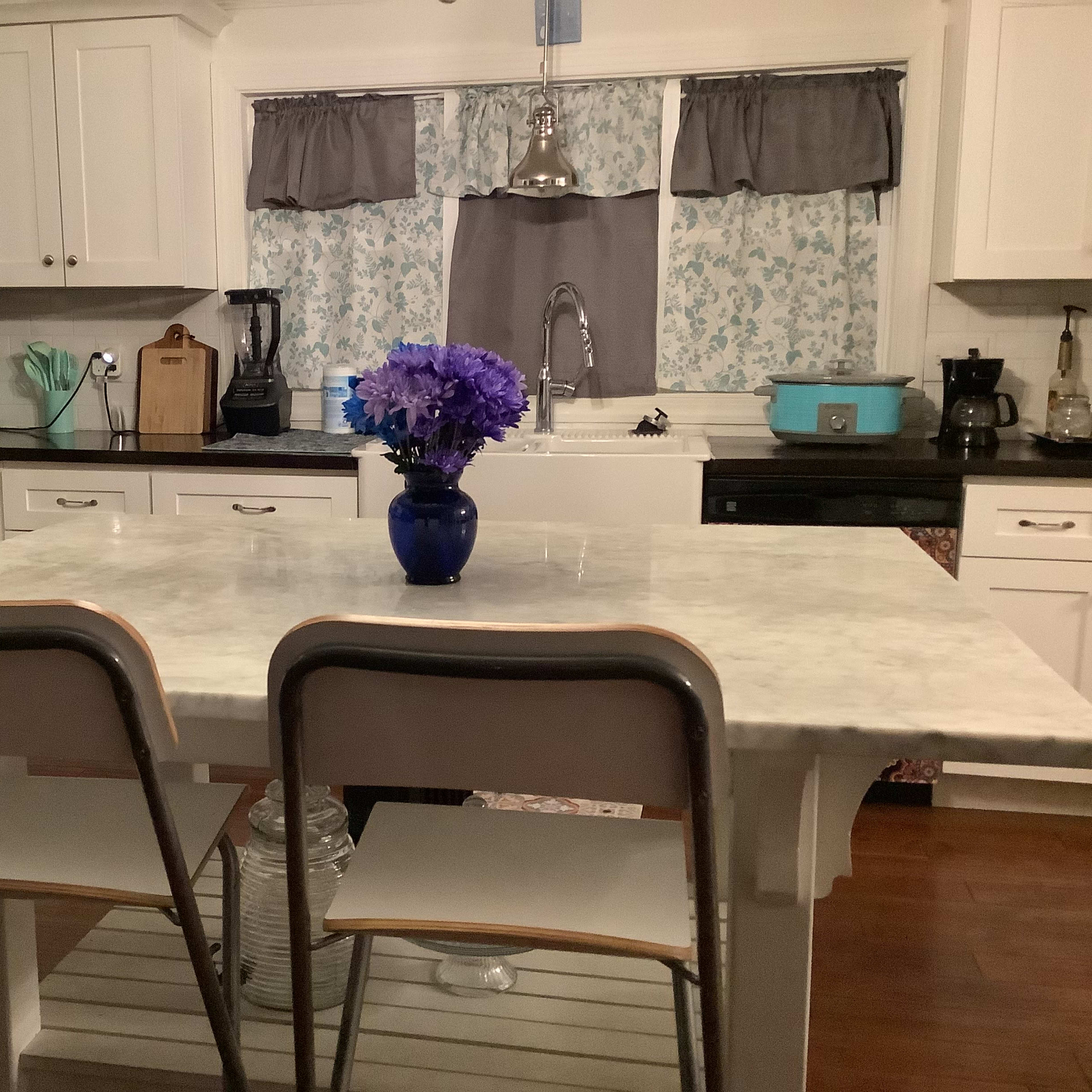 A kitchen featuring a marble-topped island with two chairs and a vase of purple flowers, surrounded by white cabinets and appliances.