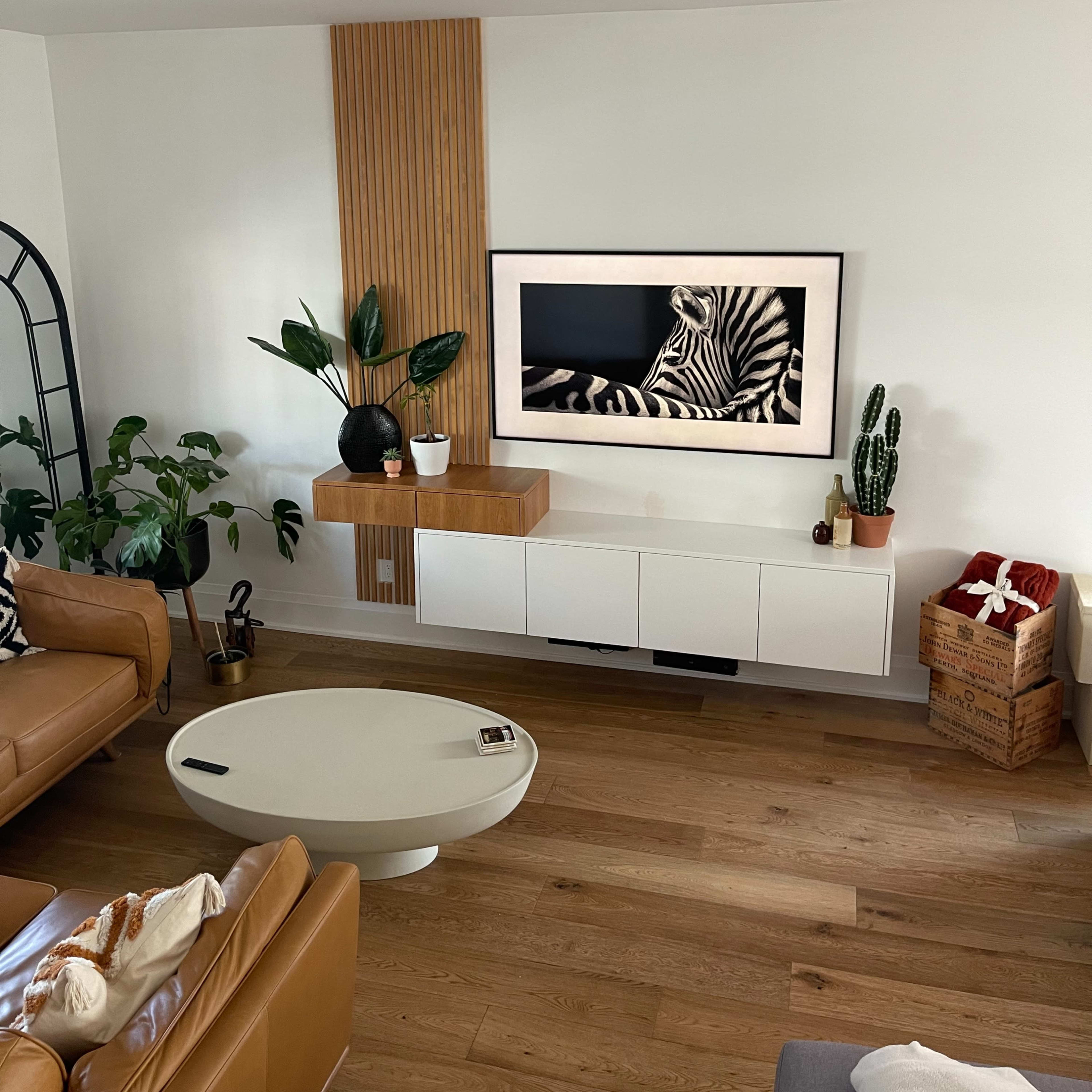 A modern living room featuring a wall-mounted television above a white cabinet, surrounded by indoor plants and a round coffee table on a wooden floor.