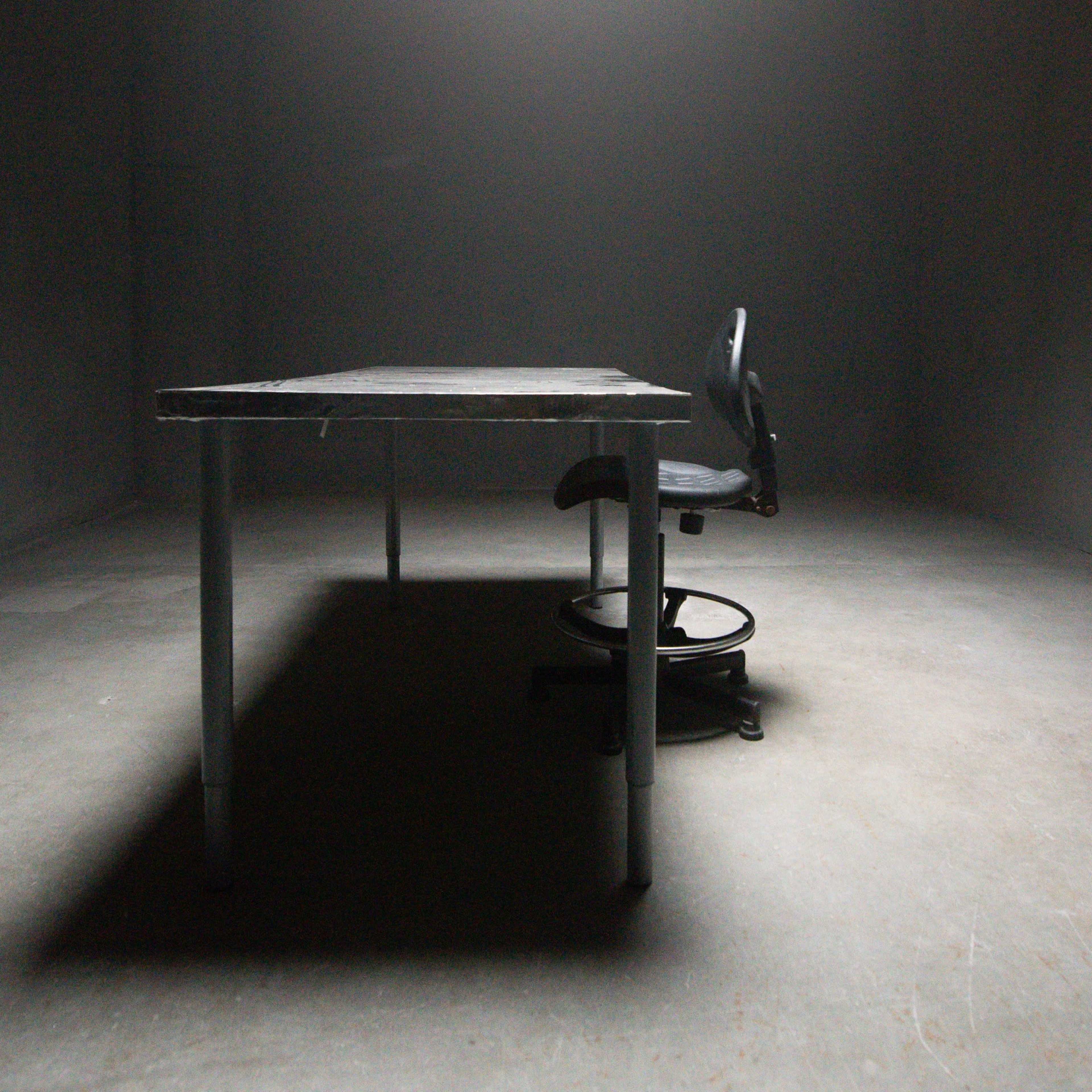 A solitary table and a chair are positioned in a dimly lit, empty room with gray walls.