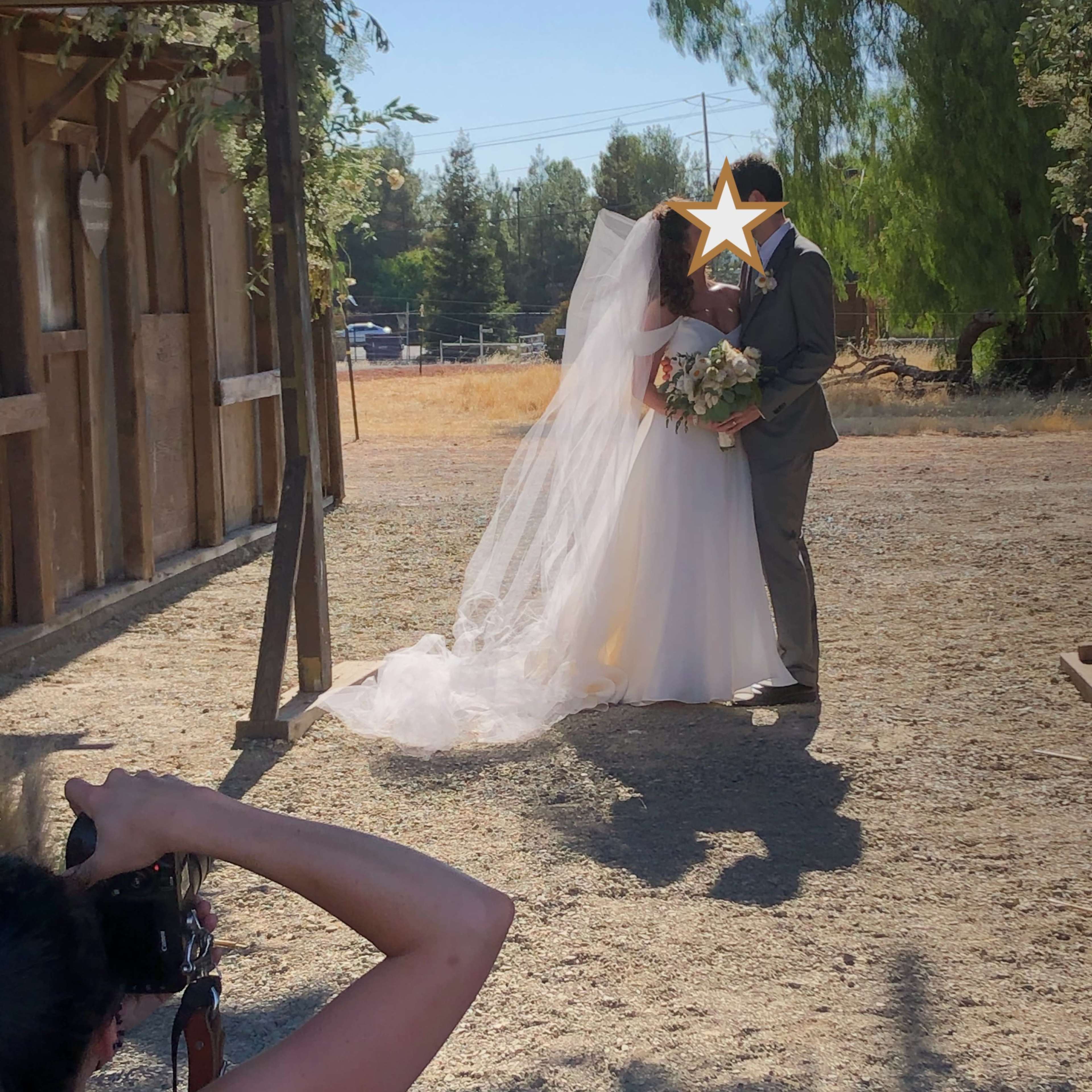 A bride and groom embrace in a sunlit outdoor setting, while a photographer captures the moment nearby.