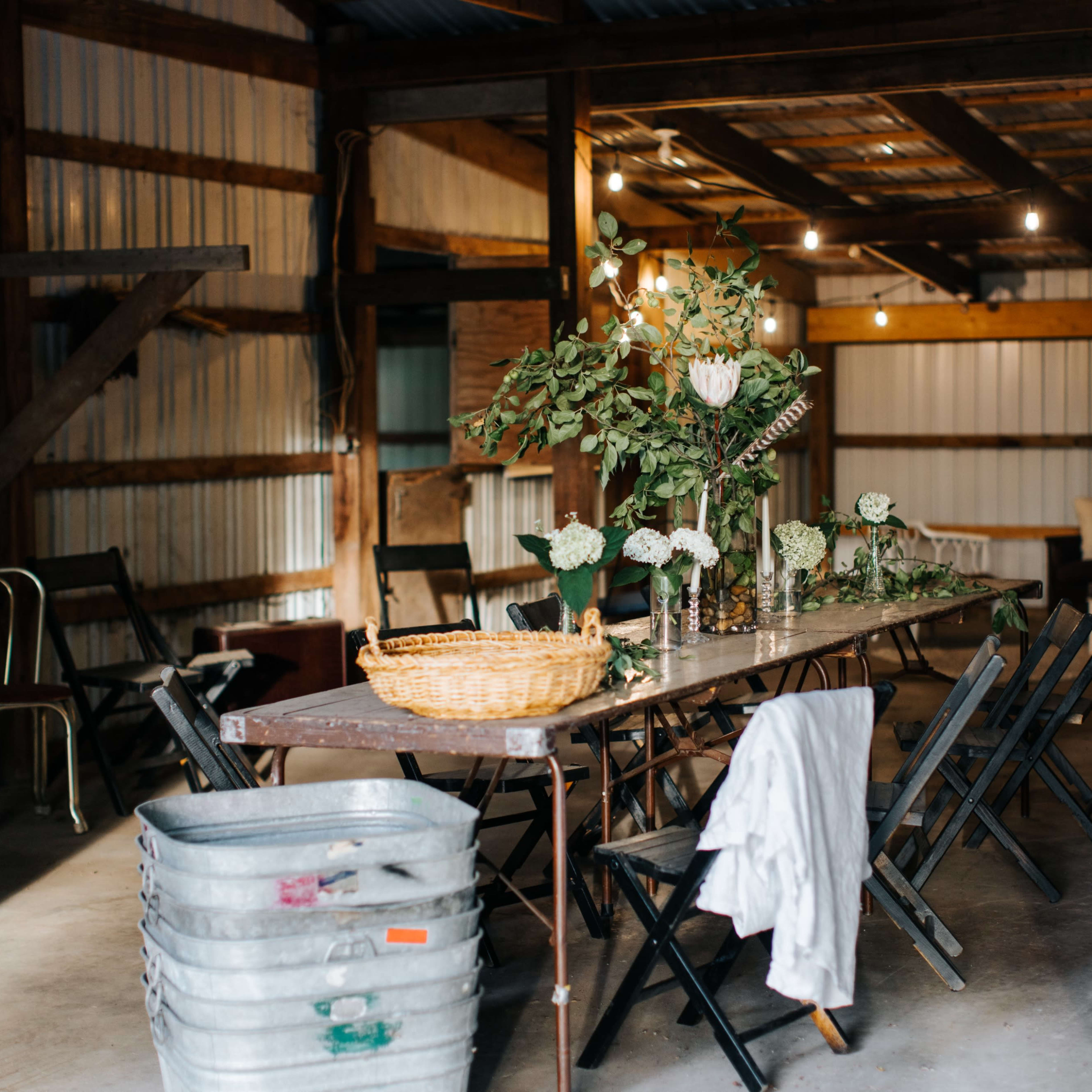 A long, wooden table is set with vases of flowers amid a rustic barn interior, featuring metal chairs and soft lighting.