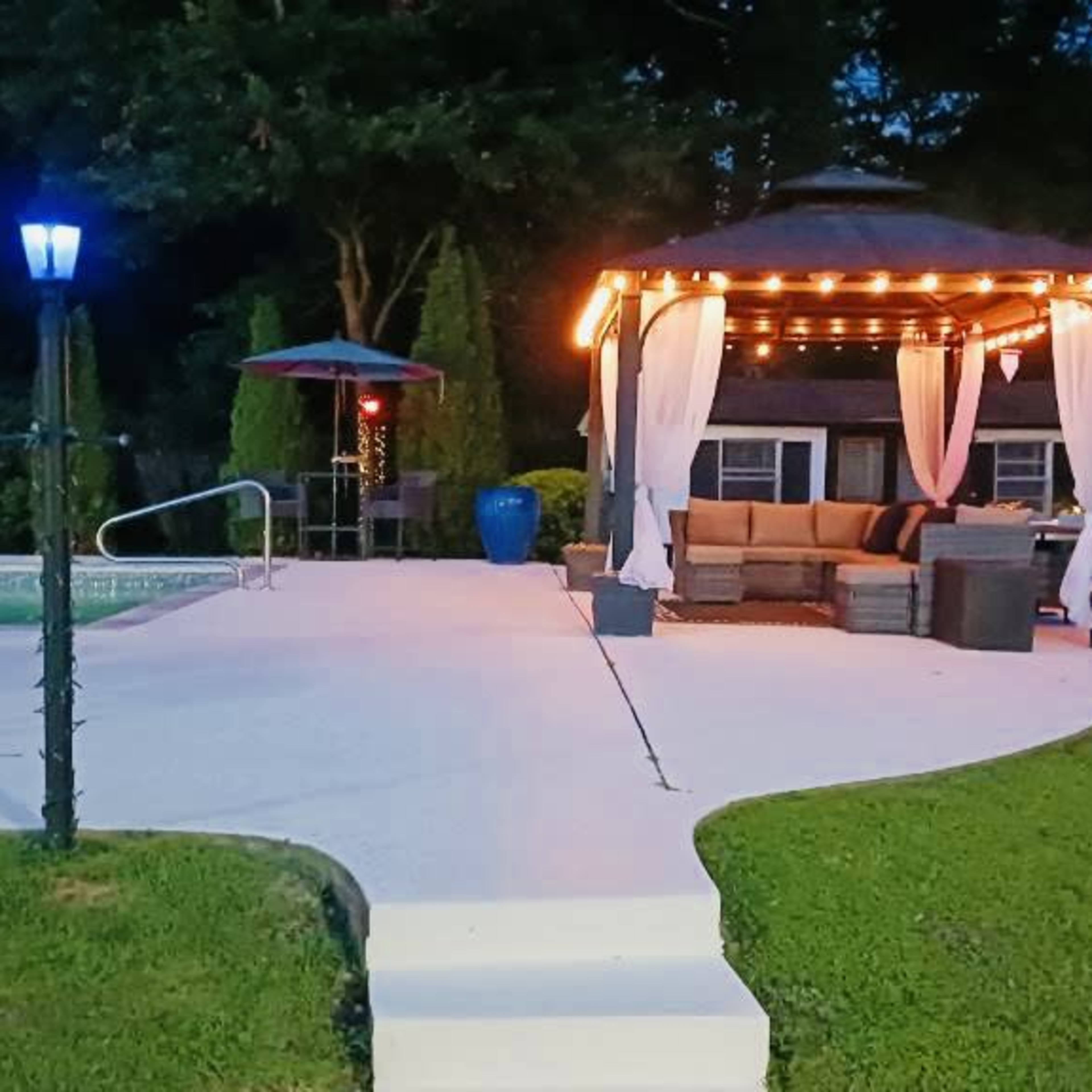 The scene features a landscaped backyard at dusk, showcasing a pool, a gazebo with string lights, and lounge chairs nearby.