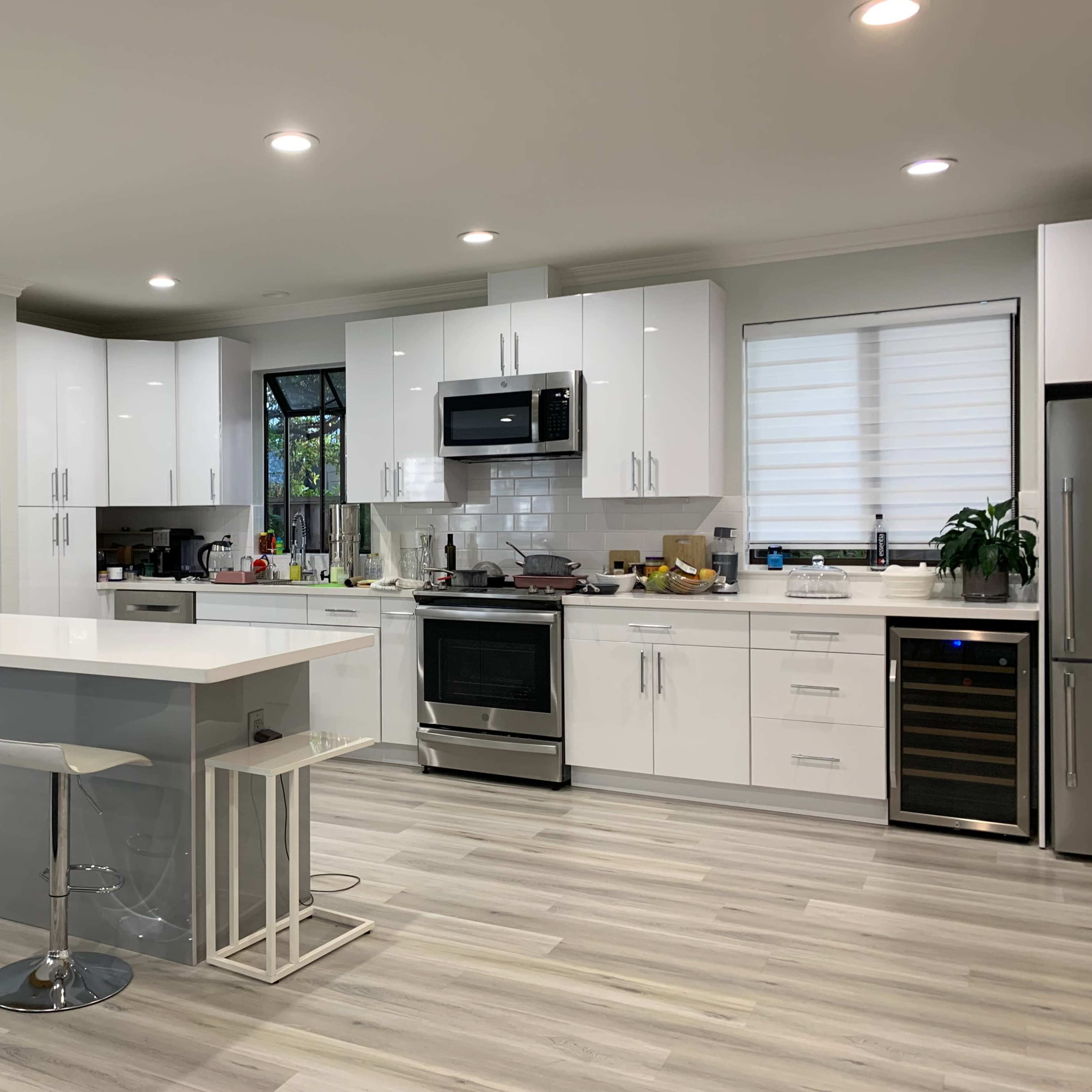 A modern kitchen with white cabinets, stainless steel appliances, a central island with bar seating, and gray wood-like flooring.