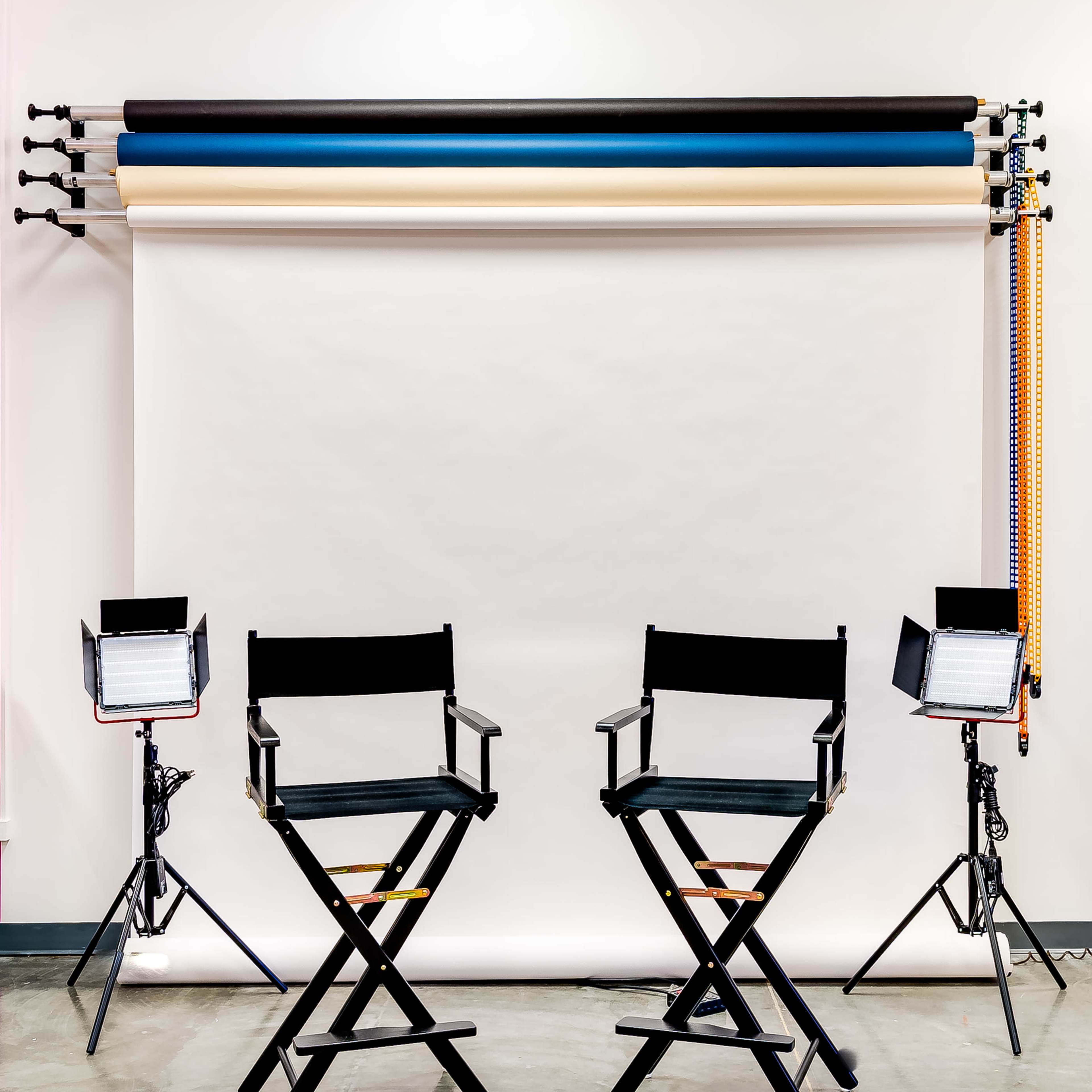 Two black director's chairs facing a plain backdrop with several rolls of colored paper and studio lights on either side.