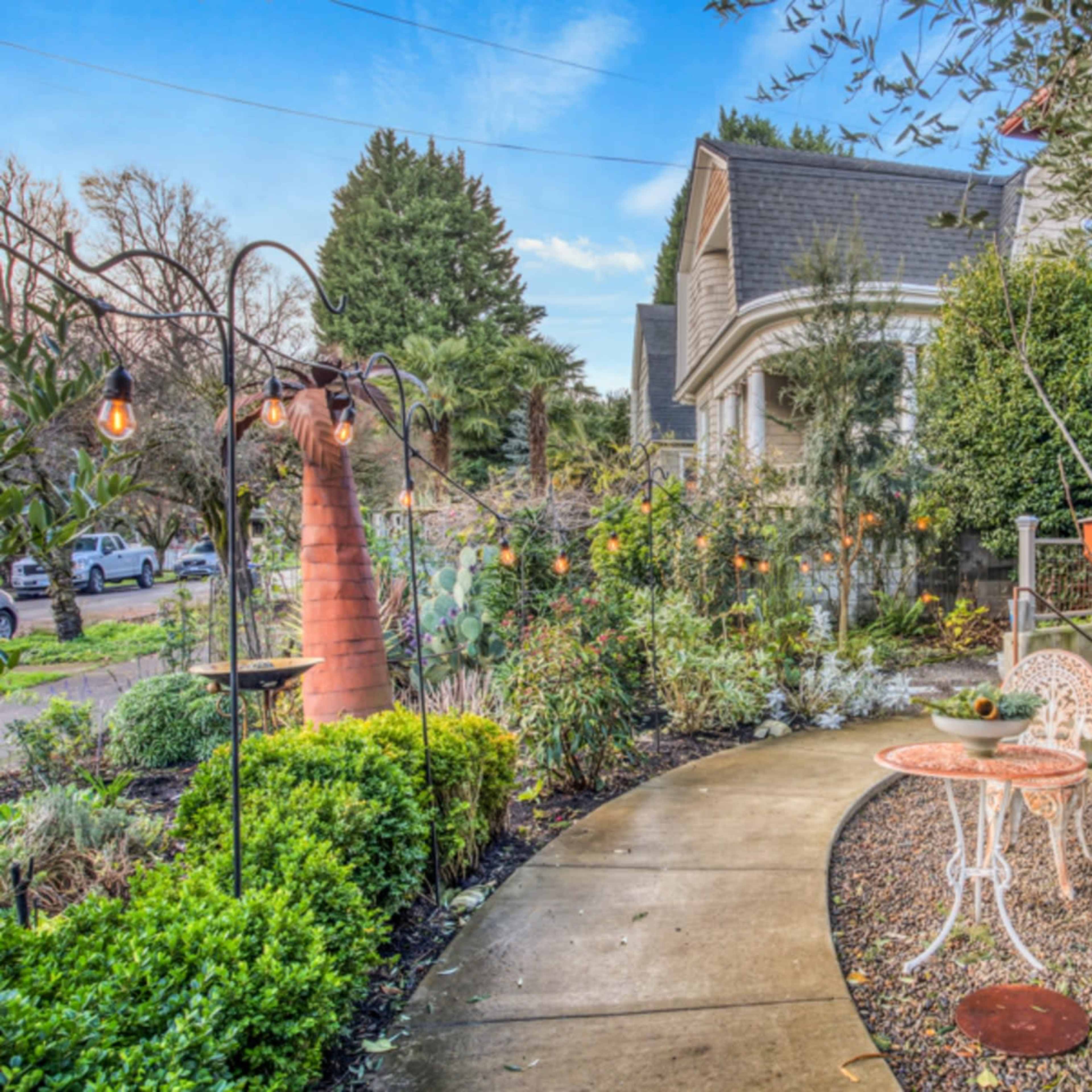 A garden path leading to a seating area with two white chairs and a small table, surrounded by various plants and decorative lighting.