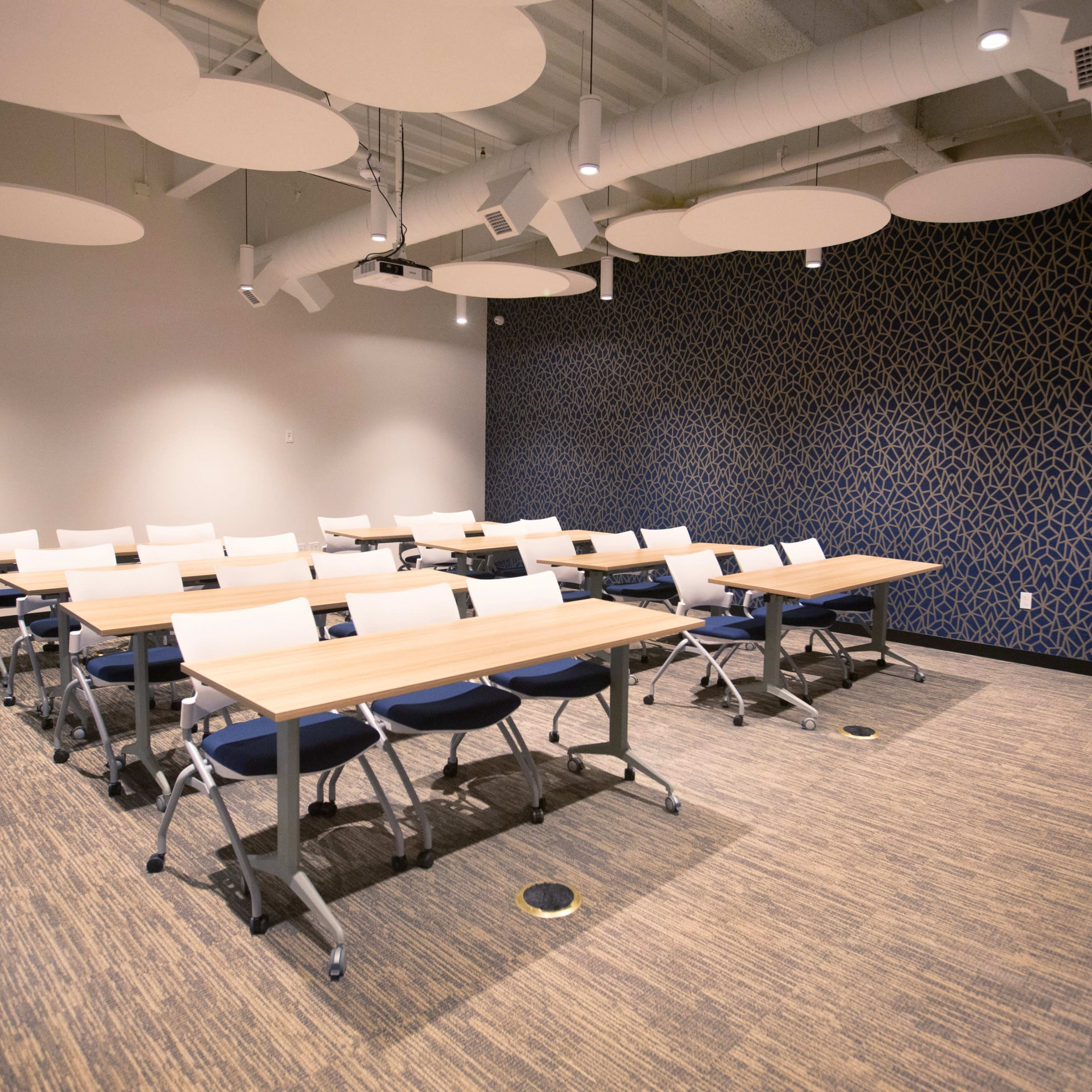 A classroom features several rows of tables and chairs, with a textured blue wall in the background and acoustic ceiling panels overhead.