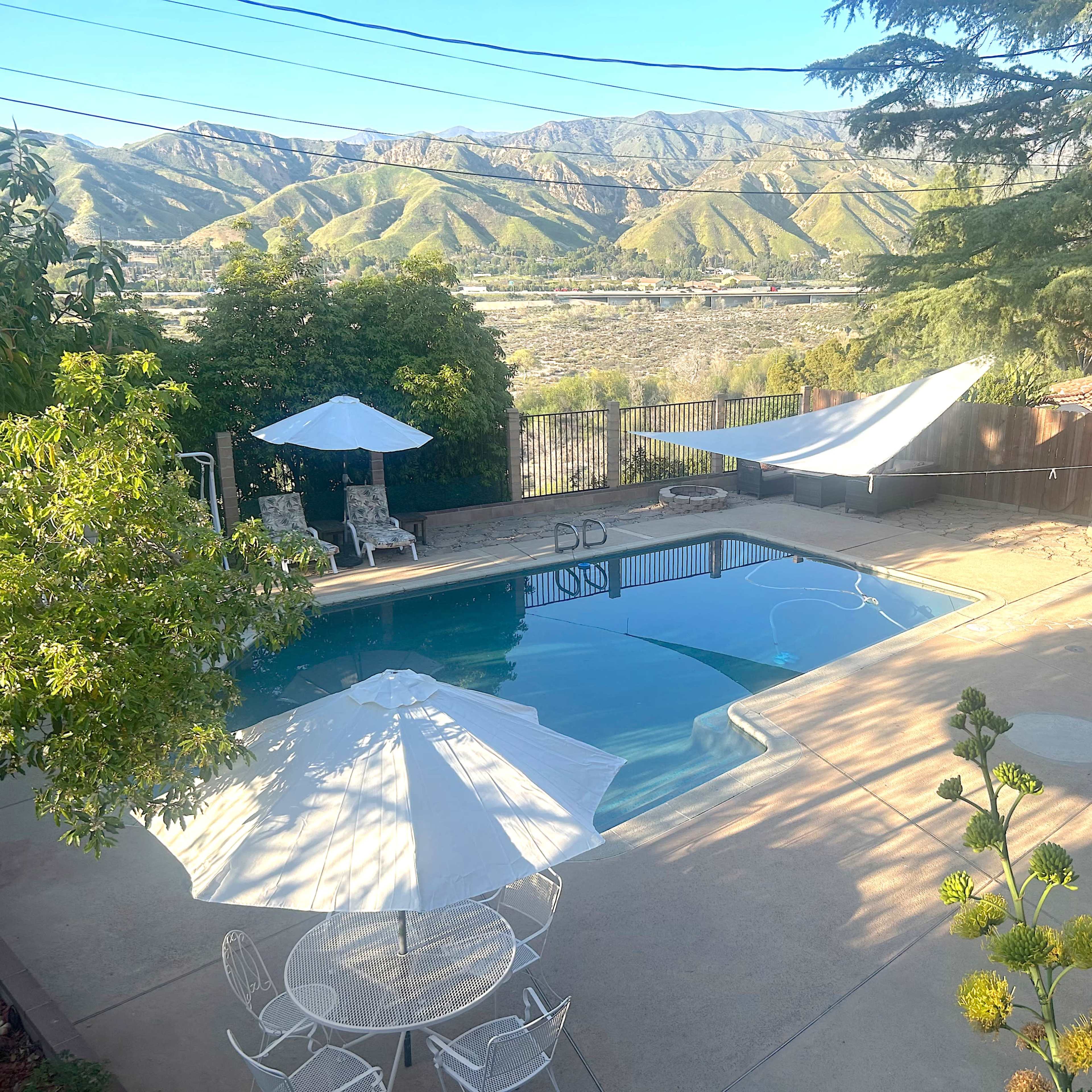A swimming pool surrounded by a concrete patio, lawn chairs, and umbrellas, with mountains visible in the background.