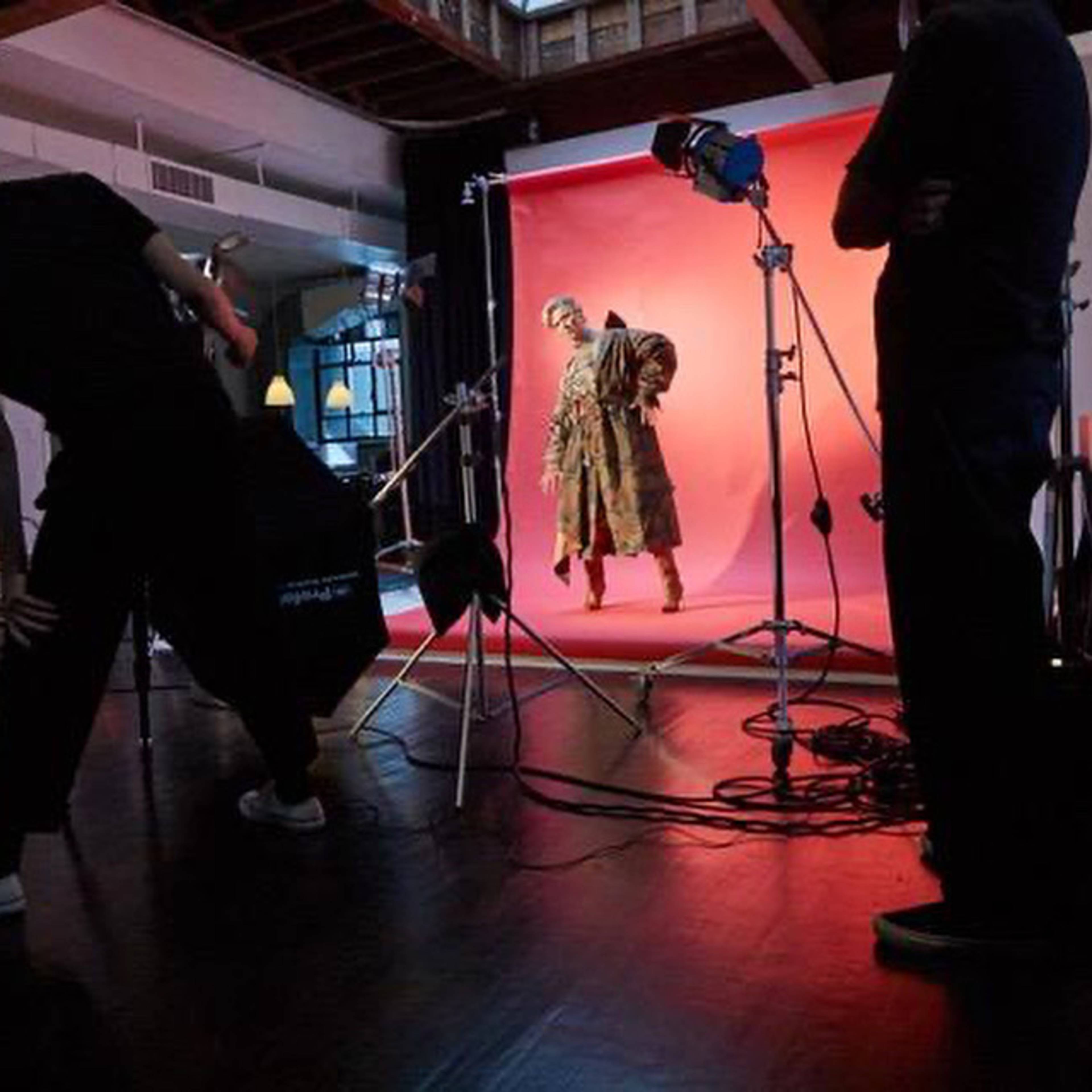 A photoshoot is taking place in a studio with a model posing against a pink backdrop while crew members adjust equipment.