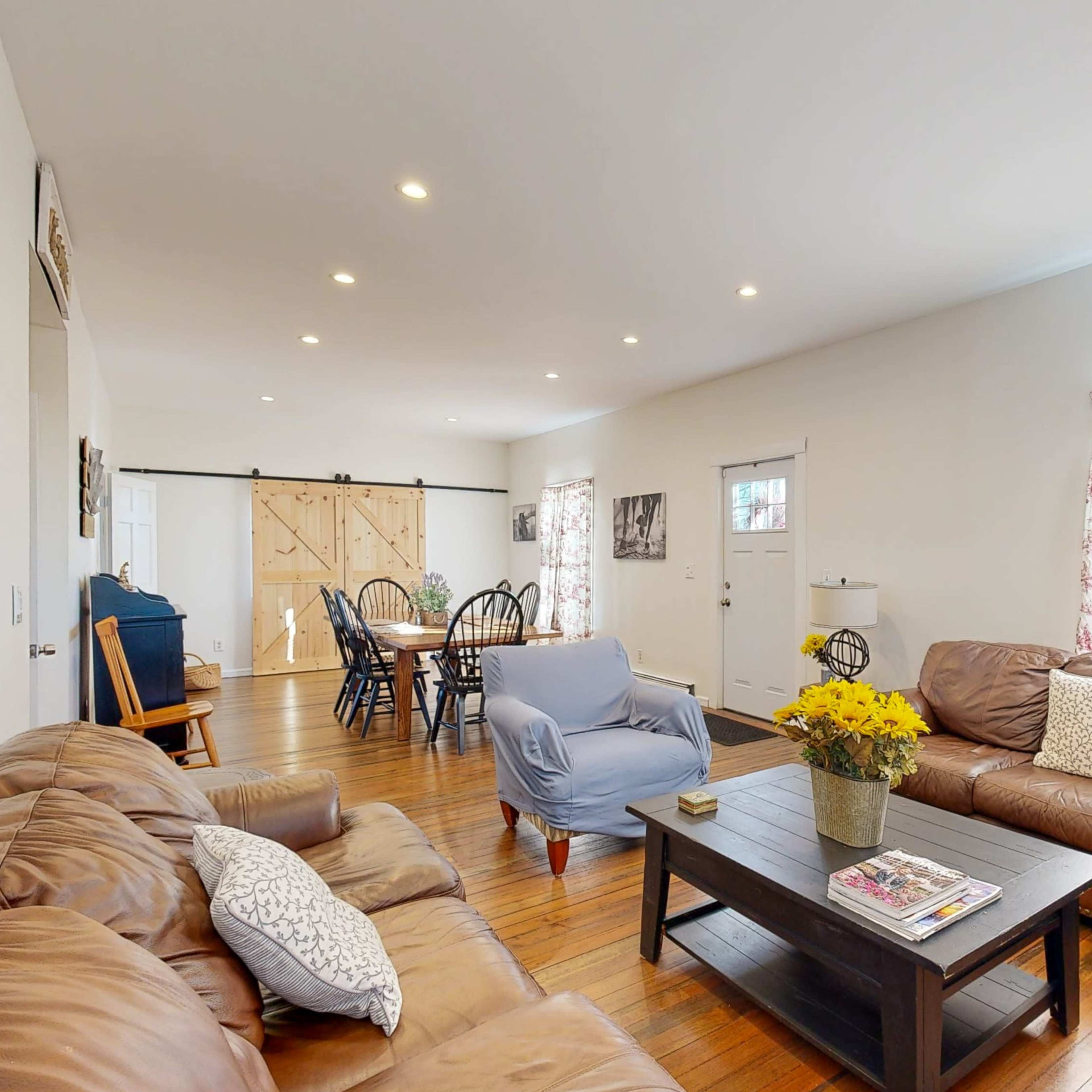 A spacious living room features leather sofas, a coffee table with magazines, and a large window with floral curtains, leading to a dining area with wooden flooring.