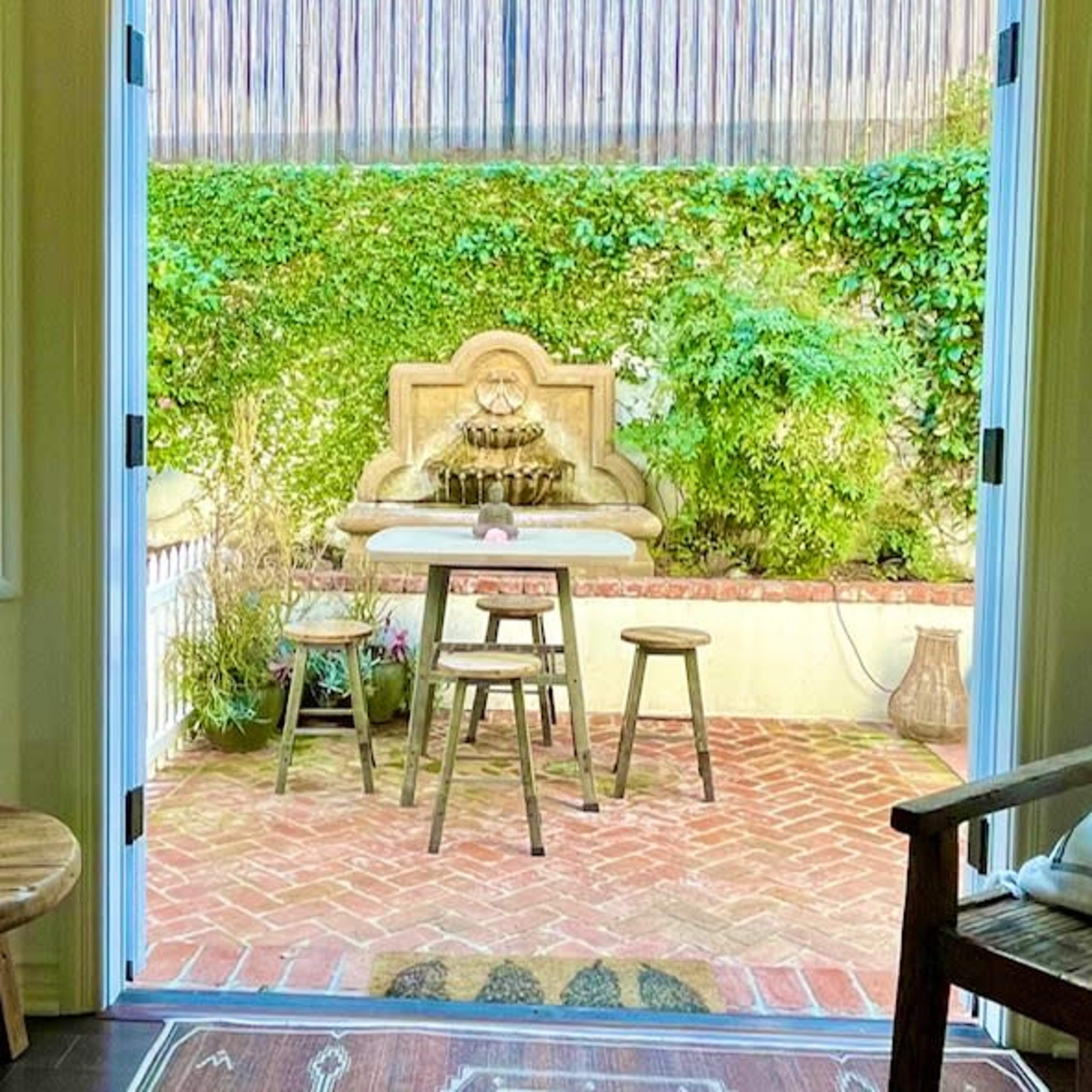 A cozy indoor space opens up to a brick-paved patio featuring a stone fountain and surrounding greenery.