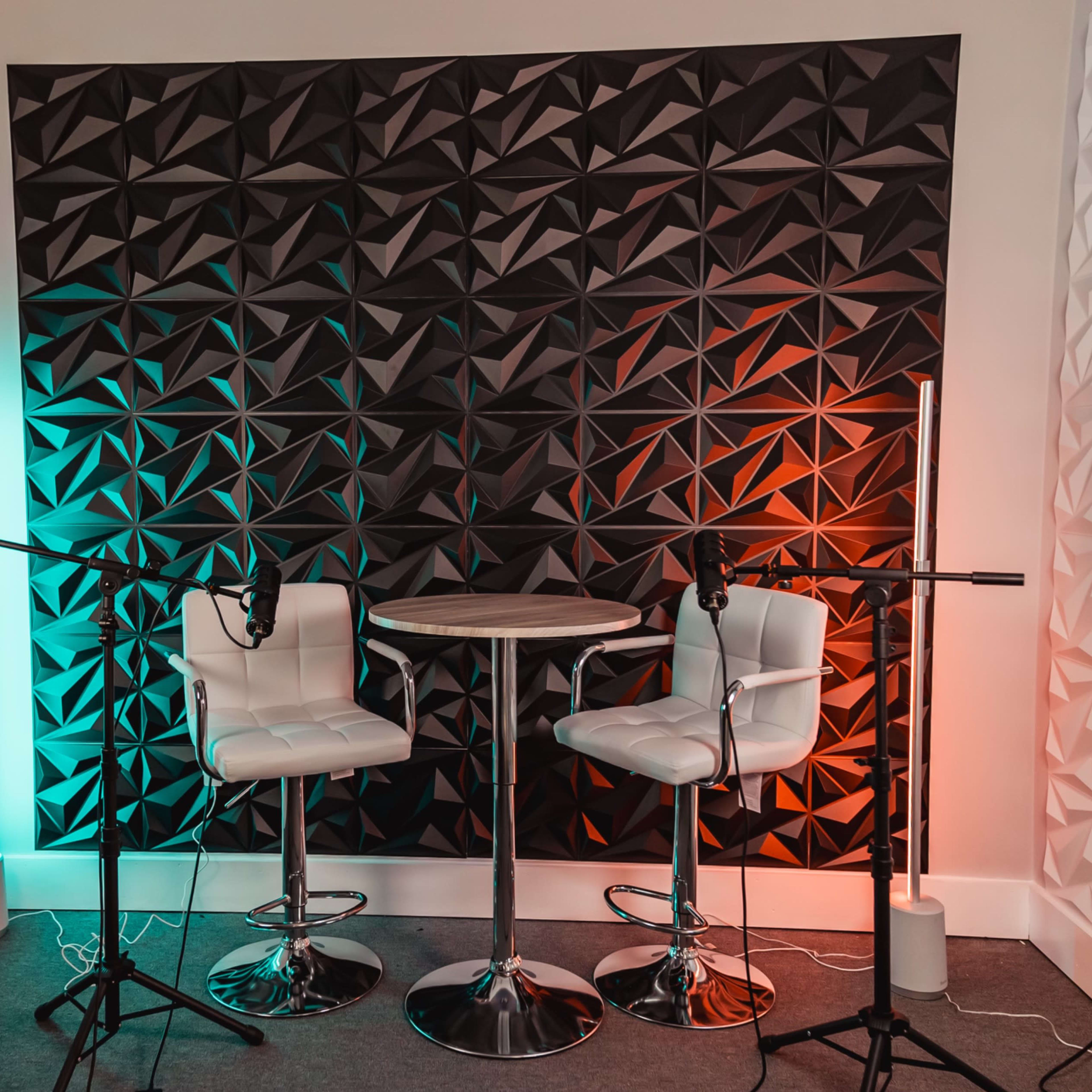 Two white bar stools positioned at a small round table in front of a textured black wall, flanked by colored light fixtures and microphones set up for recording.