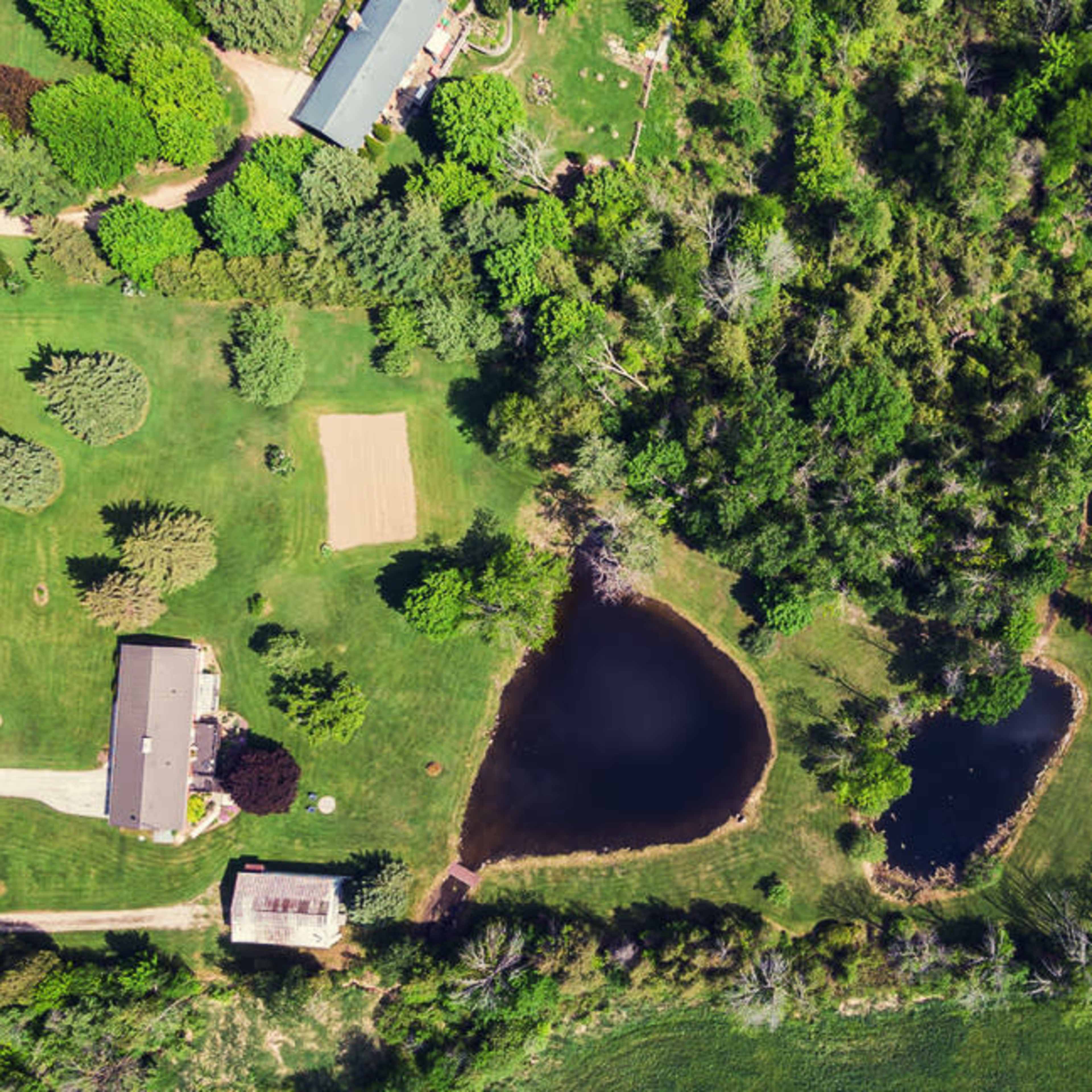 An aerial view shows a rural landscape with two ponds surrounded by greenery and several houses along a nearby road.