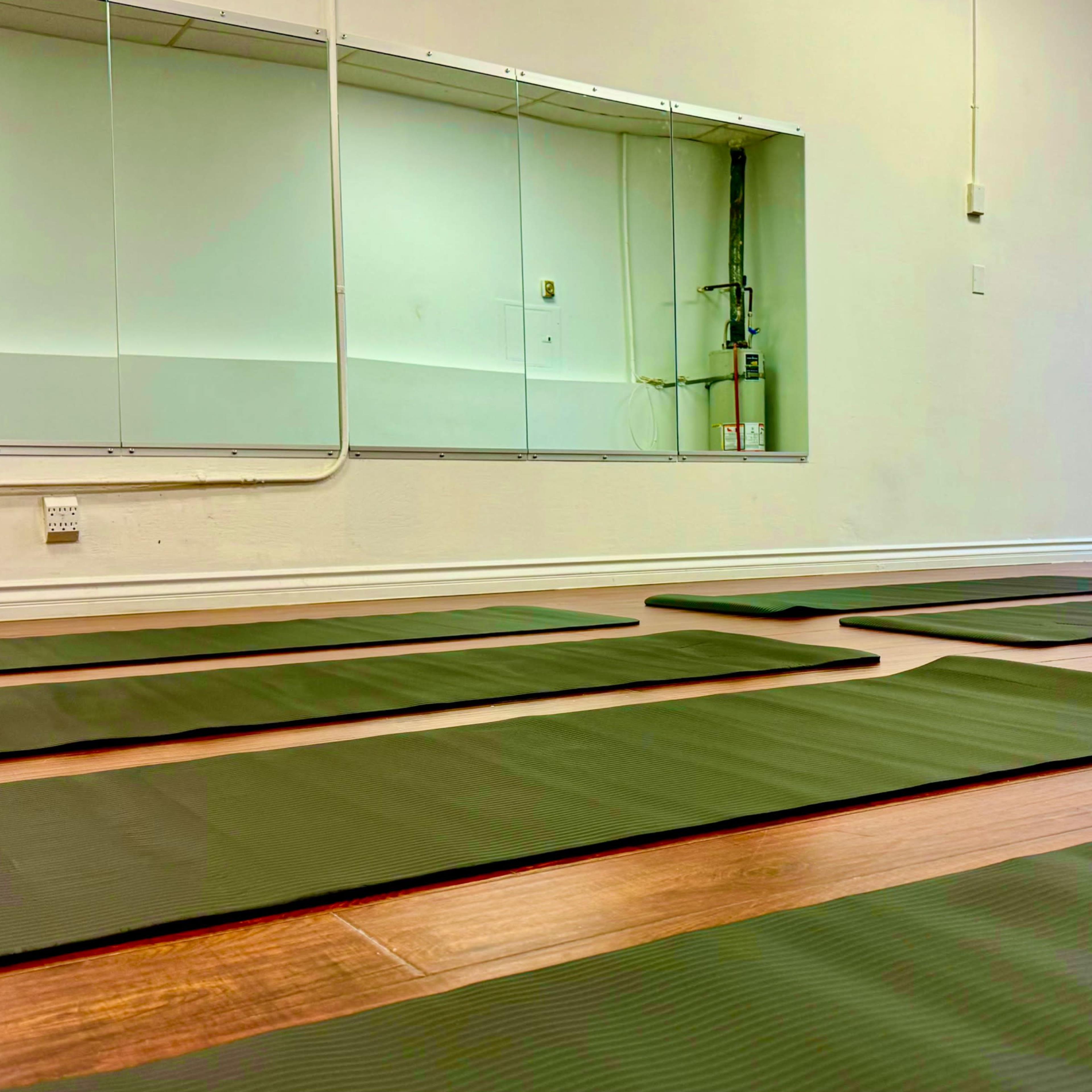 A yoga studio with several green mats arranged on a wooden floor and a mirror on the wall.