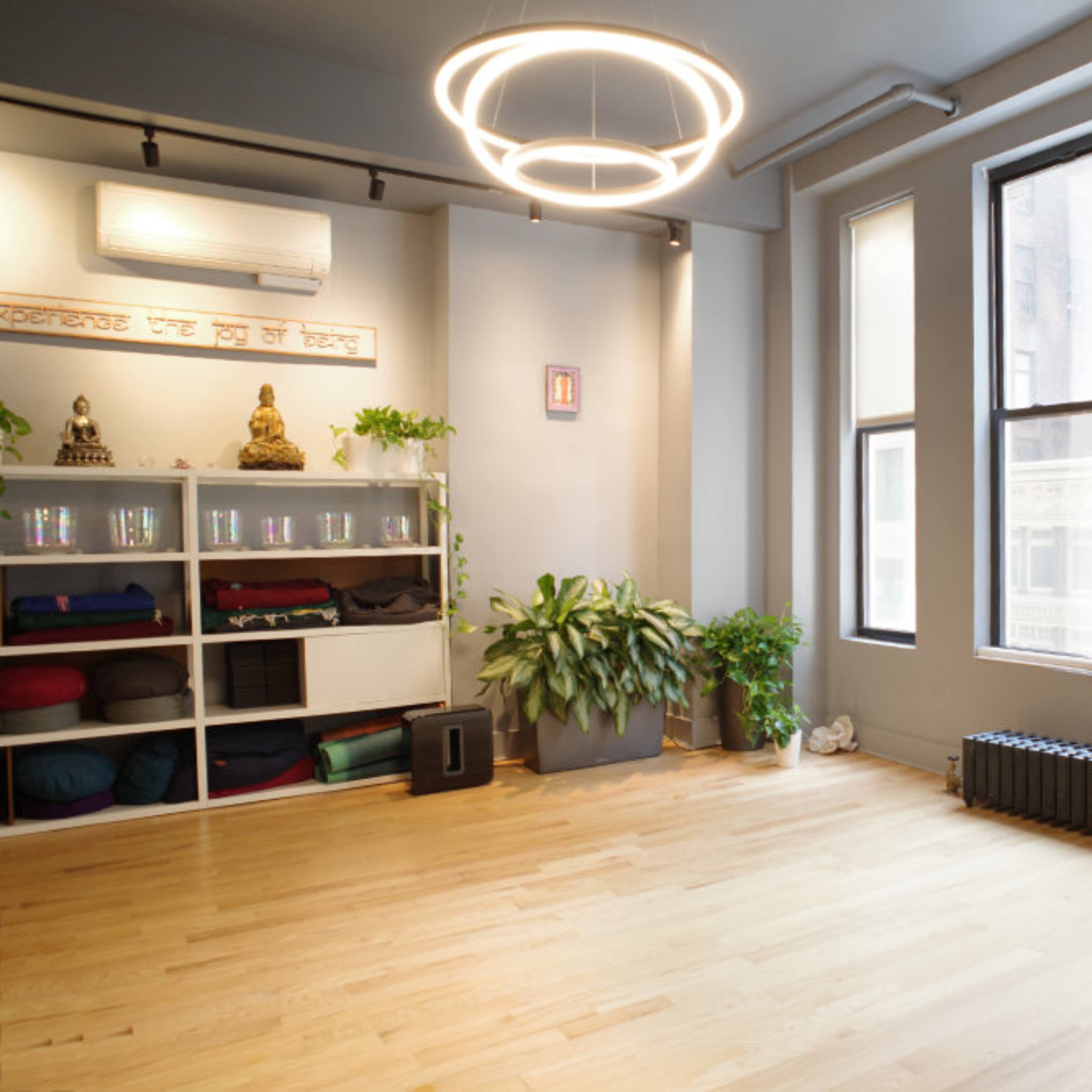 A spacious, well-lit room with wooden flooring, large windows, a modern light fixture, and shelves filled with yoga mats and equipment.