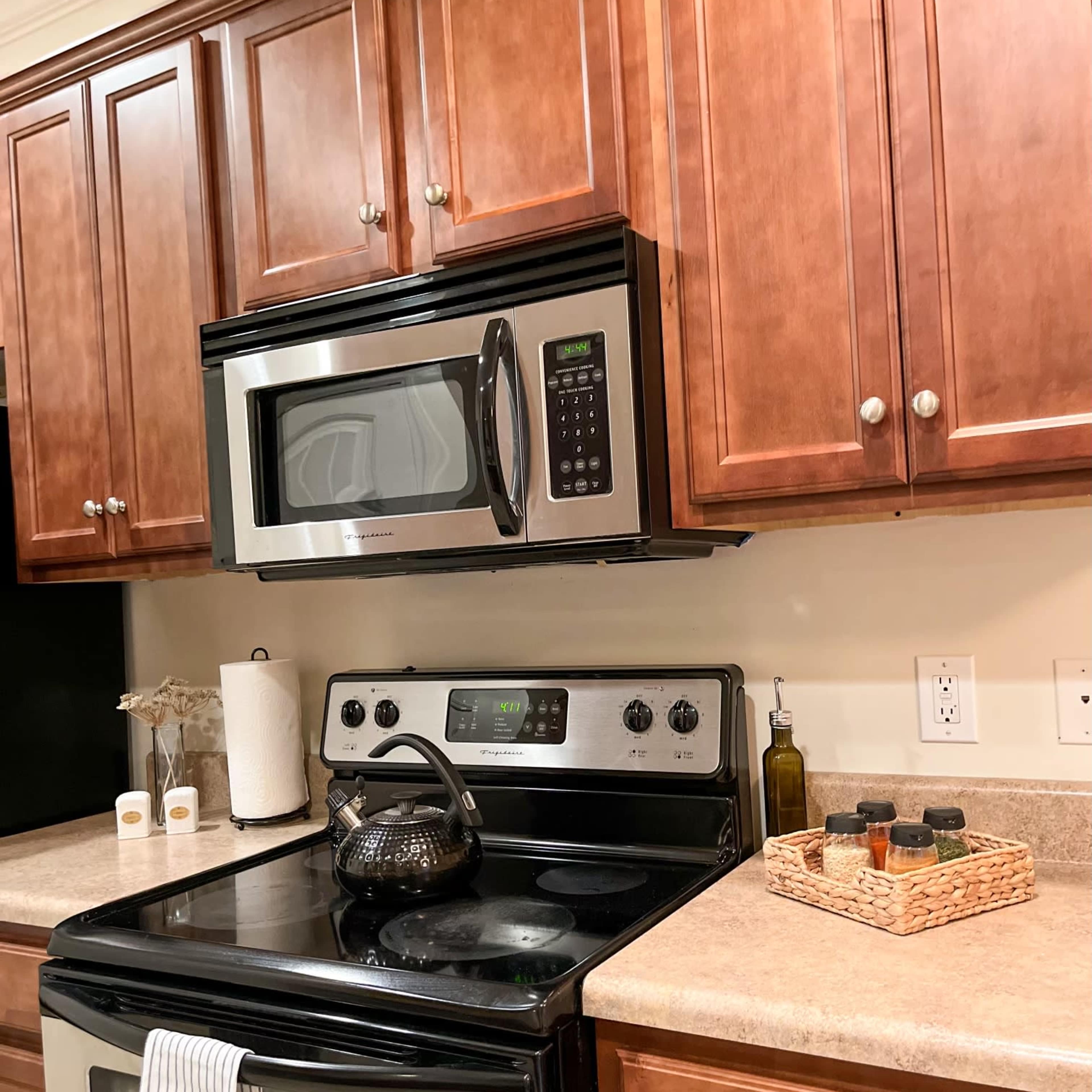A modern kitchen features wooden cabinets, a stainless steel microwave above an oven, and a stovetop with a kettle and various condiments on the countertop.