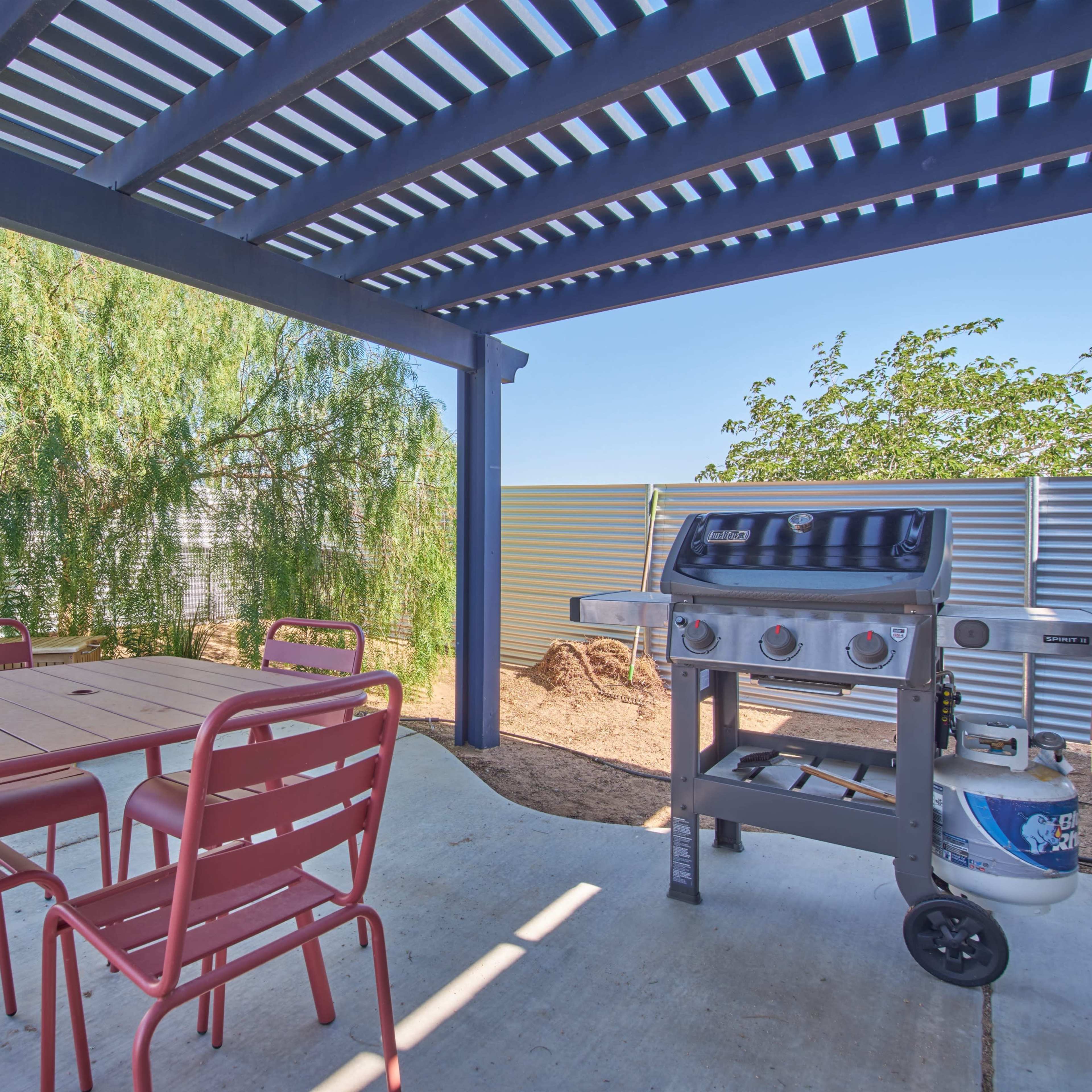 A covered patio features a table with red chairs and a gas grill beside a lush green shrub.