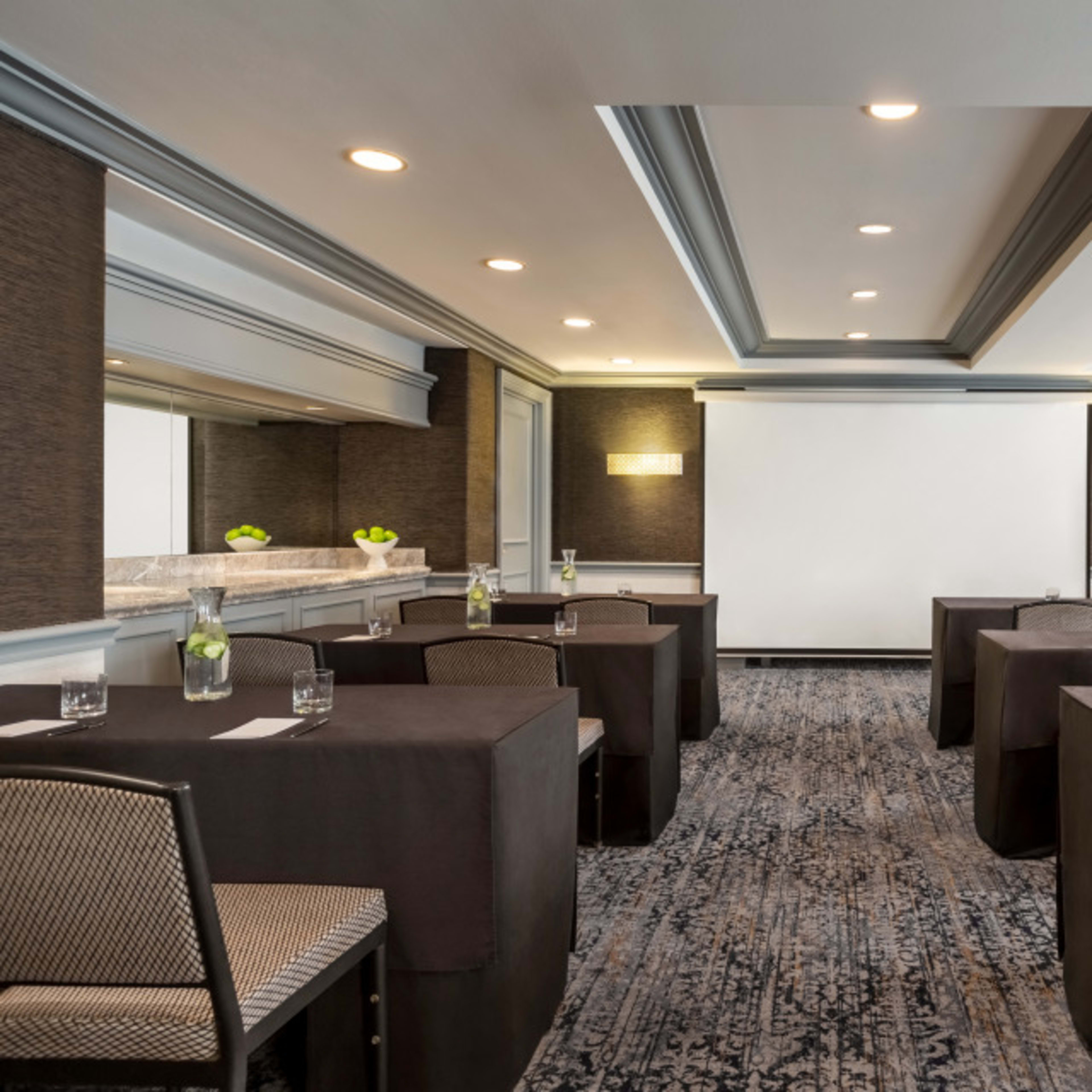A meeting room is arranged with tables covered in dark cloths, facing a large blank projection screen, with water glasses and green decorative elements on each table.