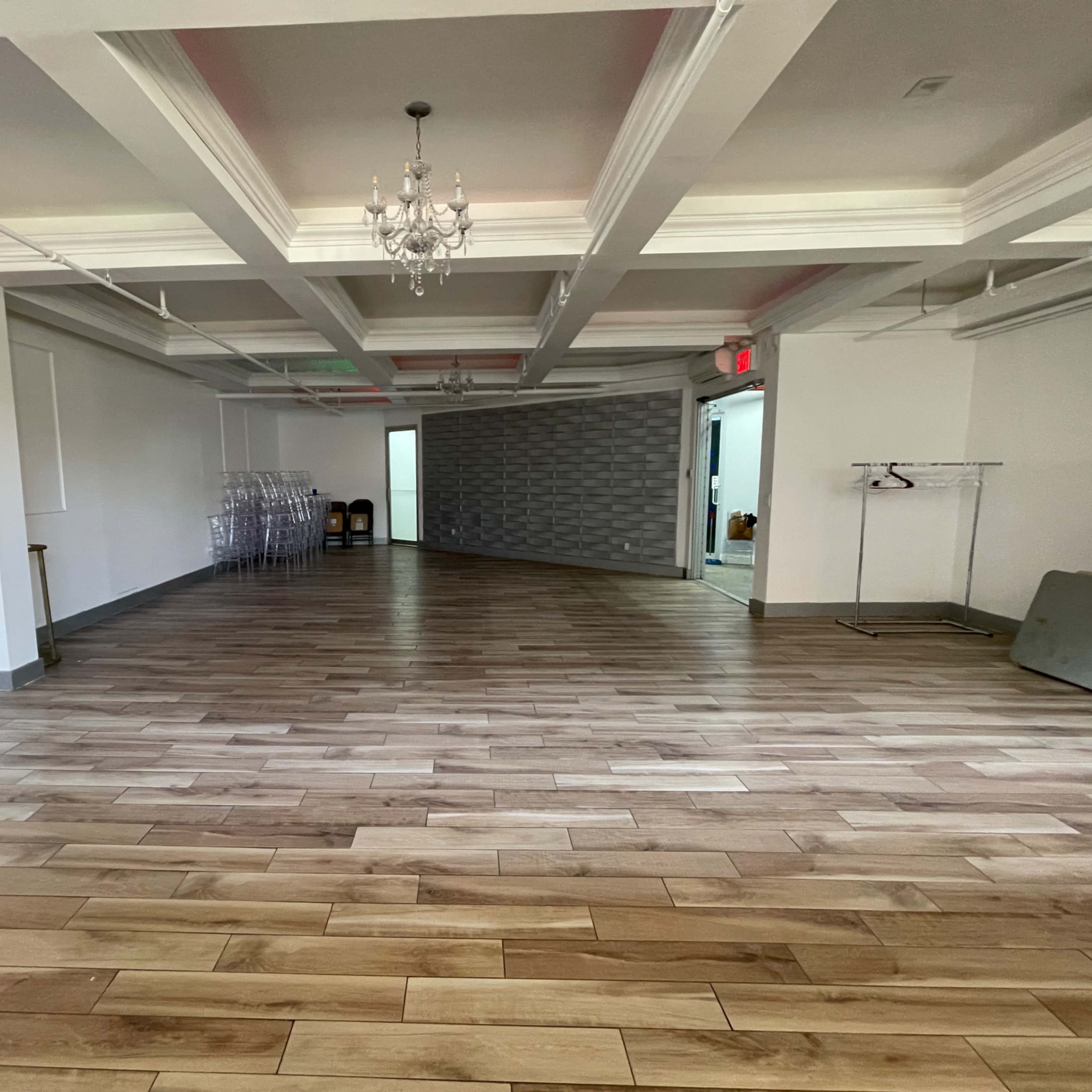 A spacious, empty room with wooden flooring and decorative ceiling beams, featuring a chandelier and exit signs.