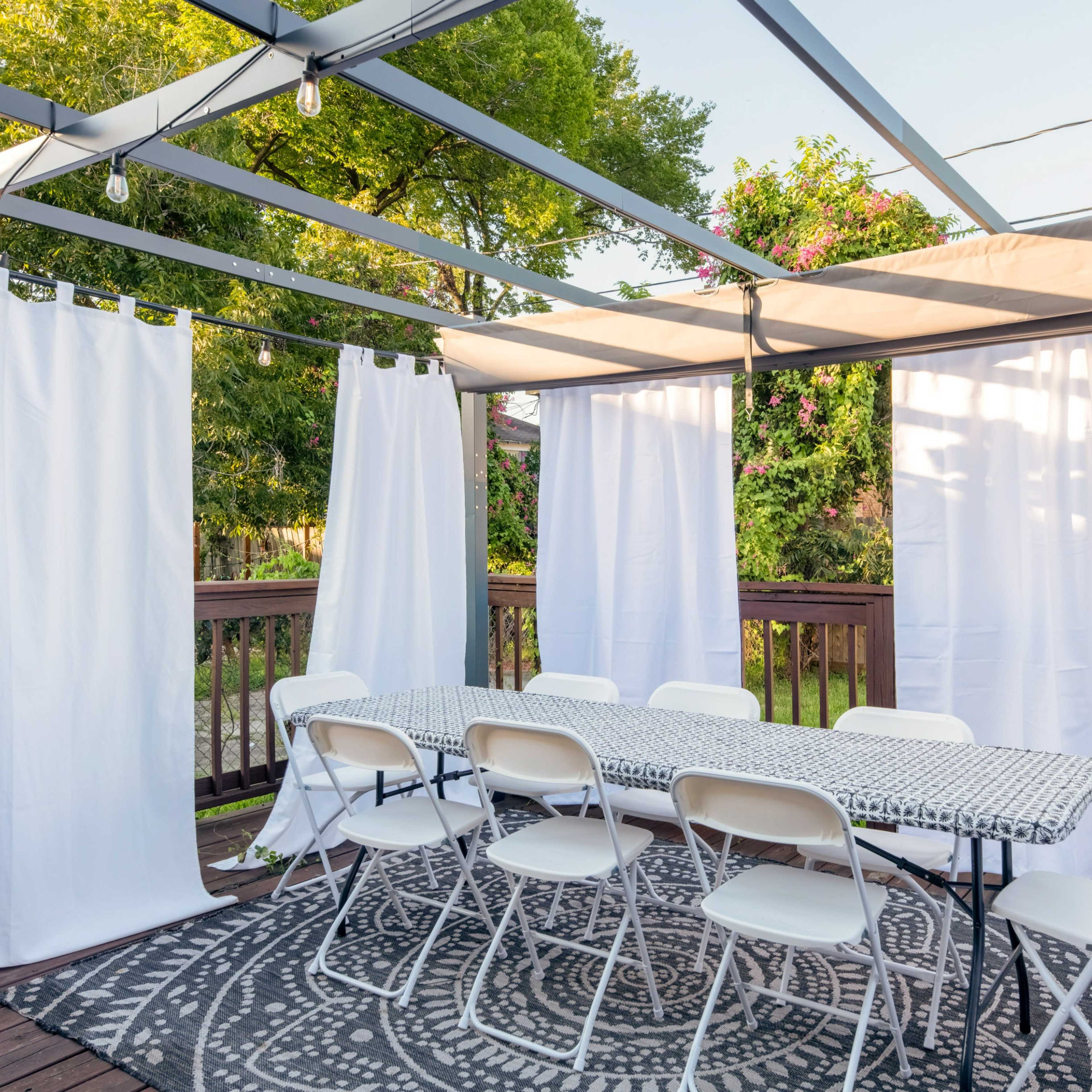 A covered outdoor dining area with white curtains, a long table set with a patterned tablecloth, and several chairs arranged around it.