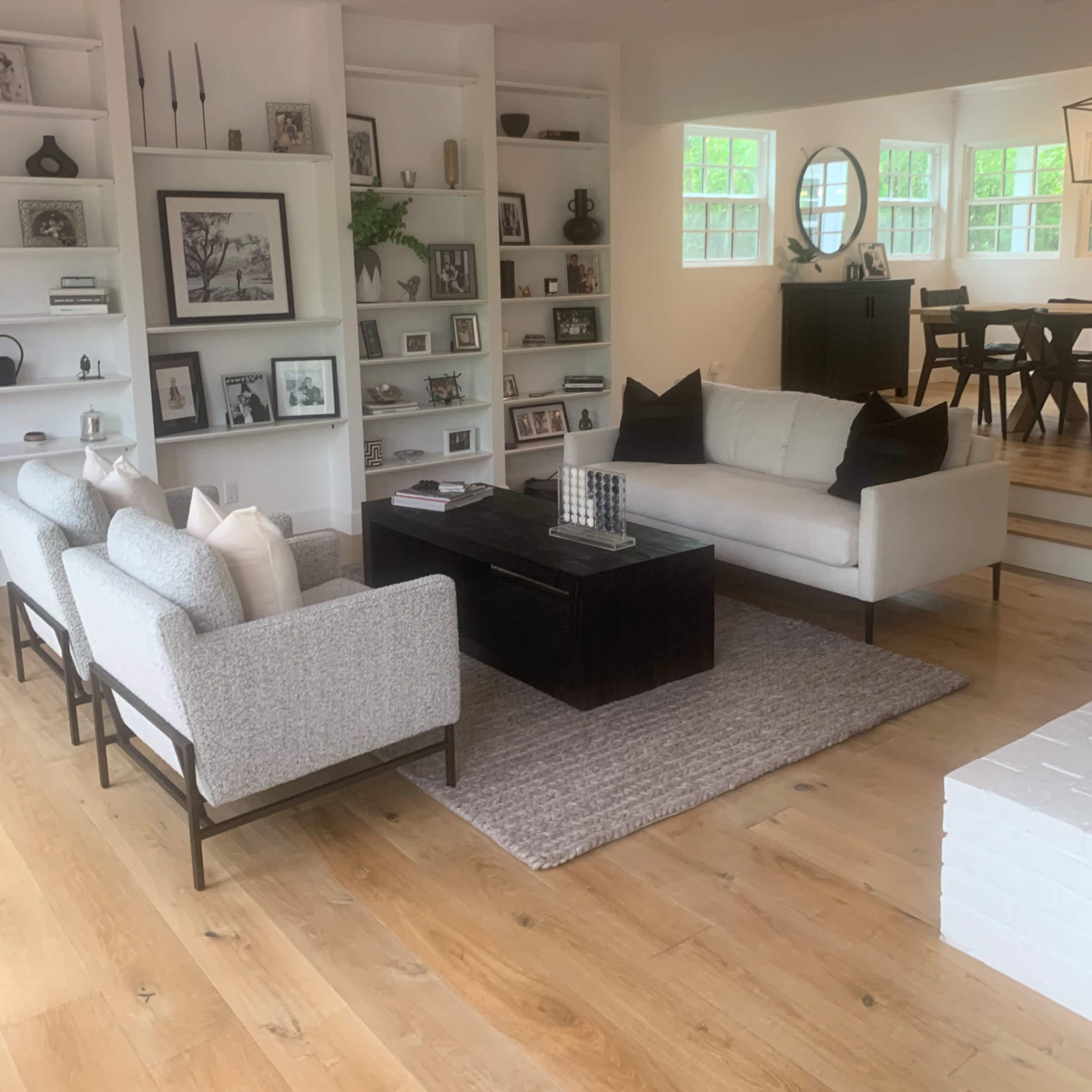A living room with a sofa and two armchairs around a coffee table, surrounded by built-in shelves filled with decorative items, and a dining area visible in the background.