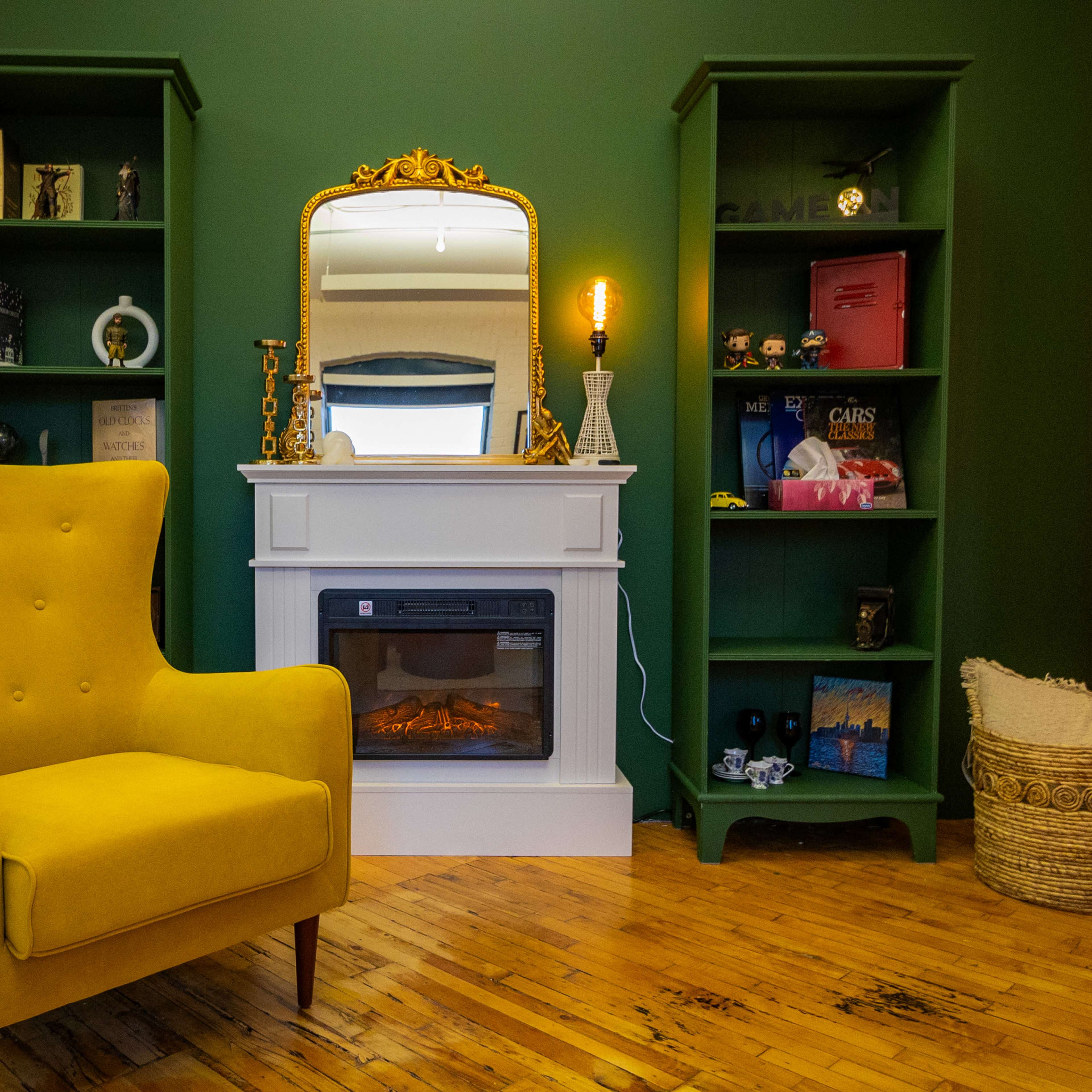 A cozy room with a yellow armchair next to a white fireplace, flanked by green bookshelves, against a deep green wall.