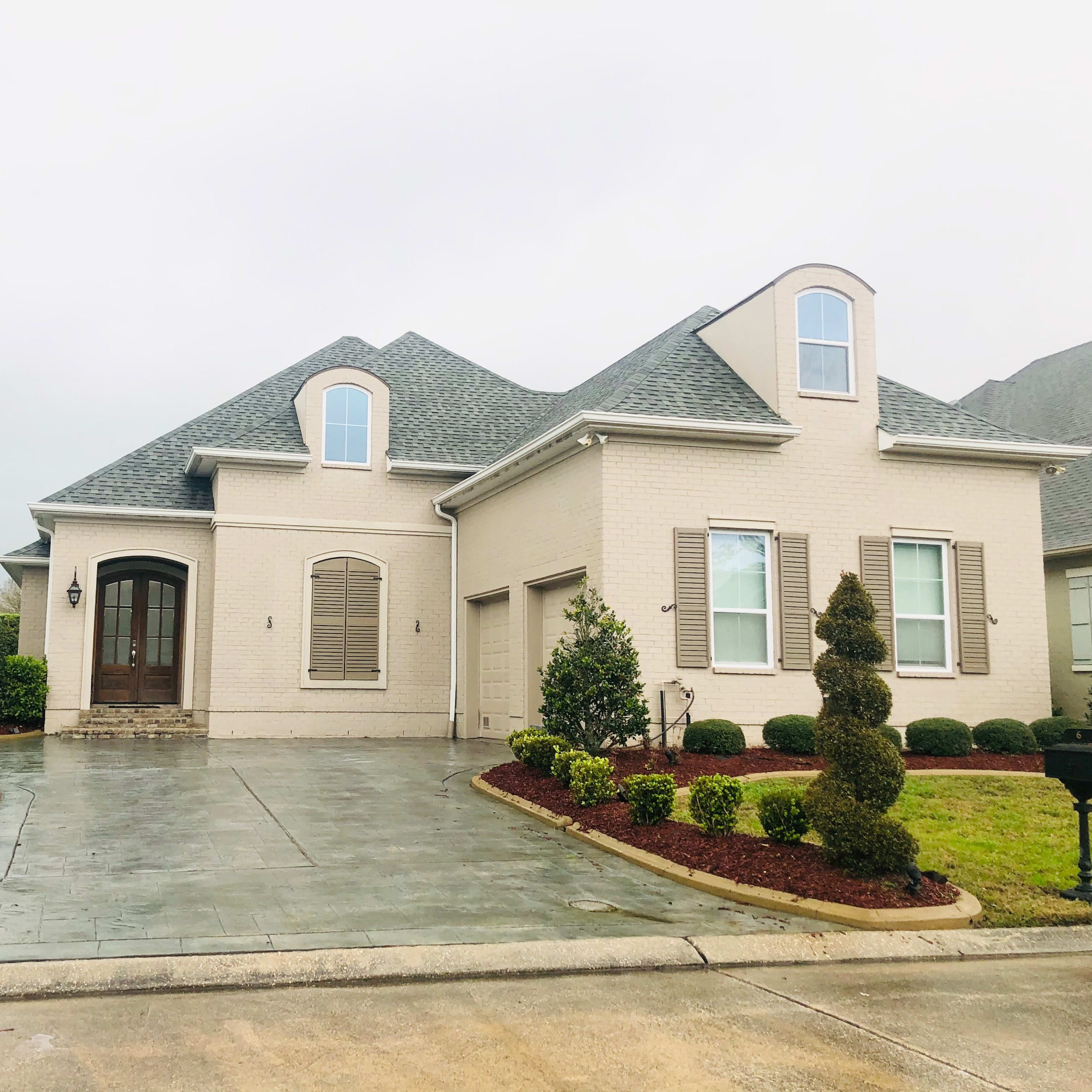 A well-maintained single-story house with a sloped roof, bordered by neatly trimmed shrubs and a paved driveway.