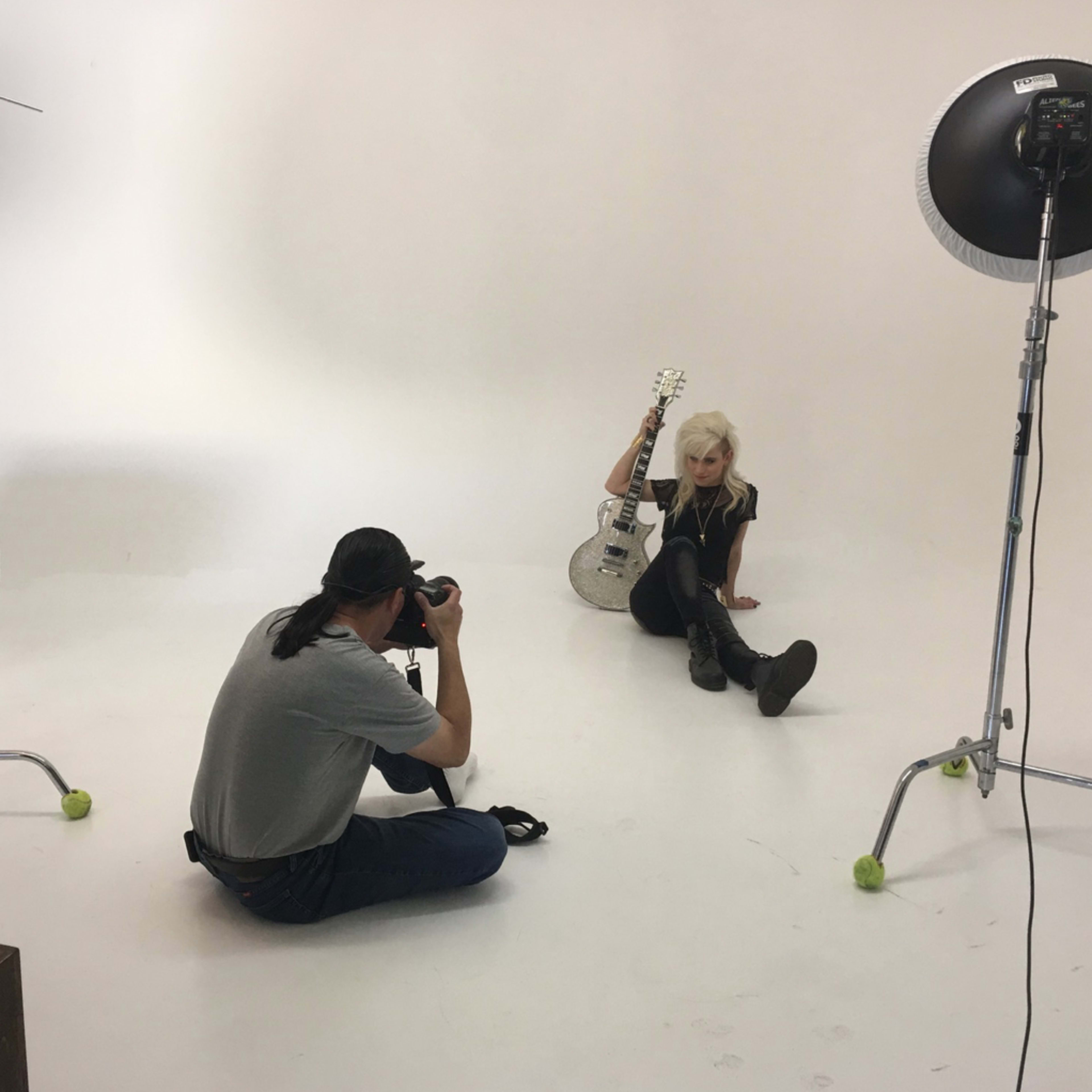 A photographer is taking pictures of a model holding a guitar in a studio with a plain white backdrop.