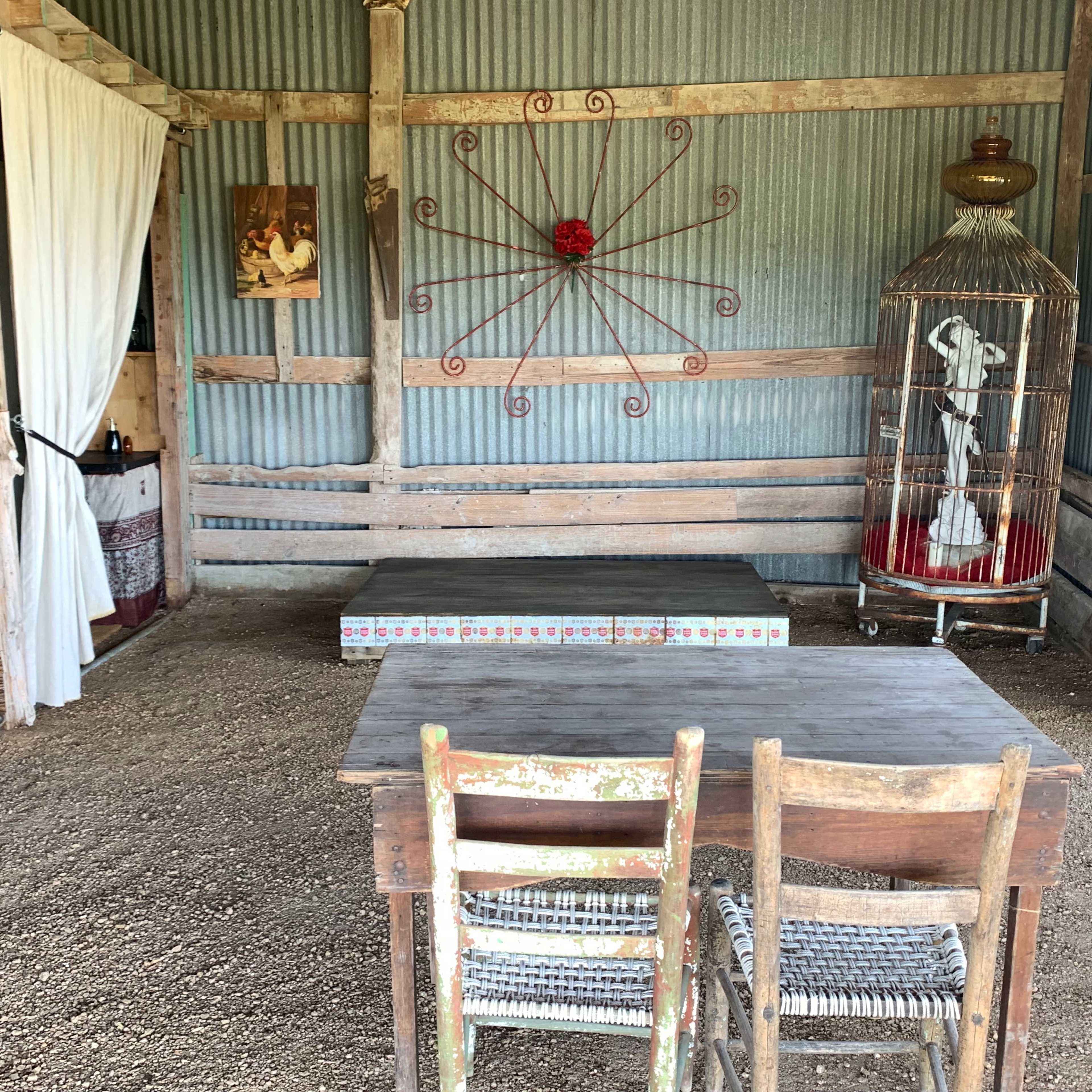 An interior space with wooden walls, a small table, two wooden chairs, and a decorative birdcage against a backdrop of corrugated metal.