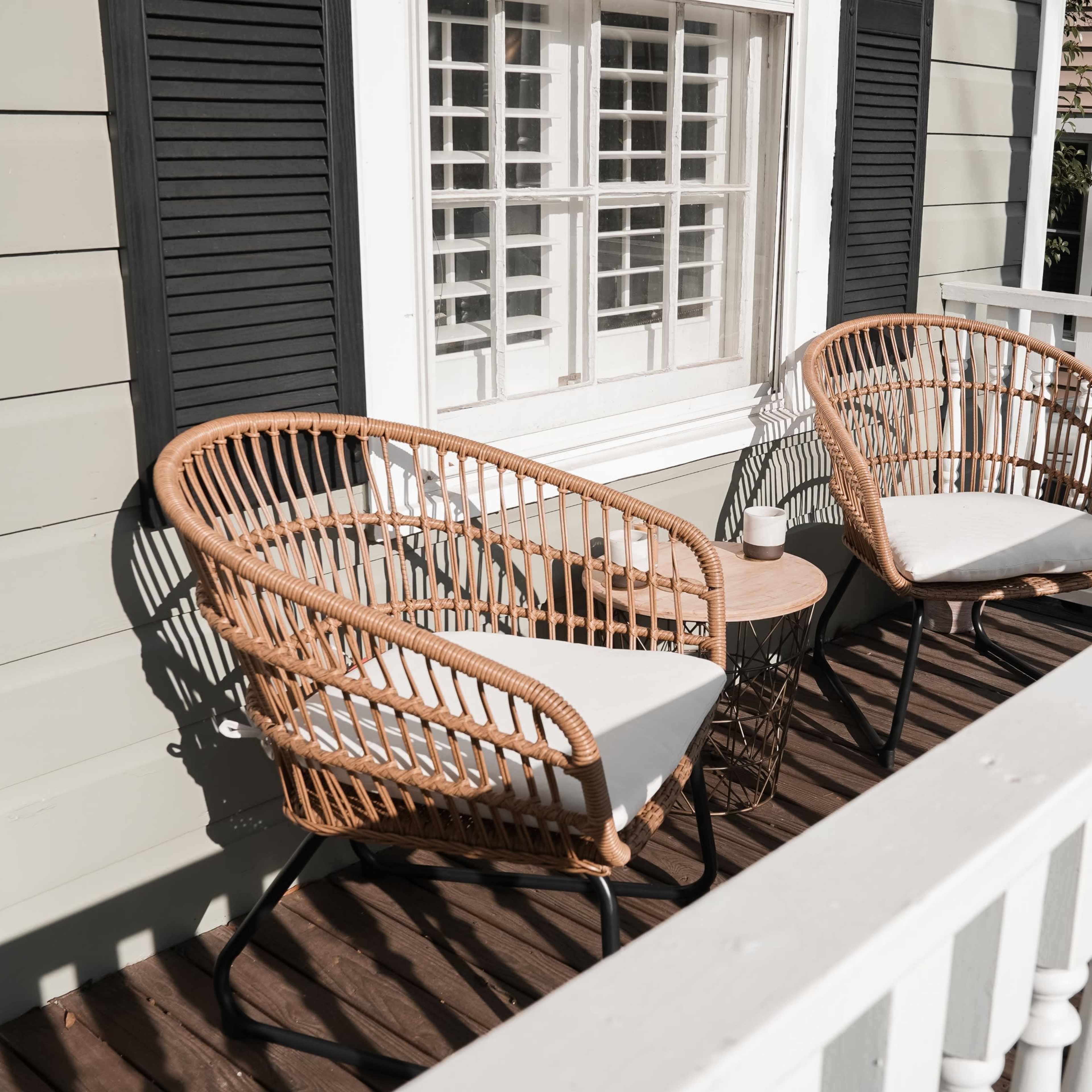 Two wicker chairs with cushions are positioned beside a small table on a balcony overlooking a house with white railings and wooden floorboards.