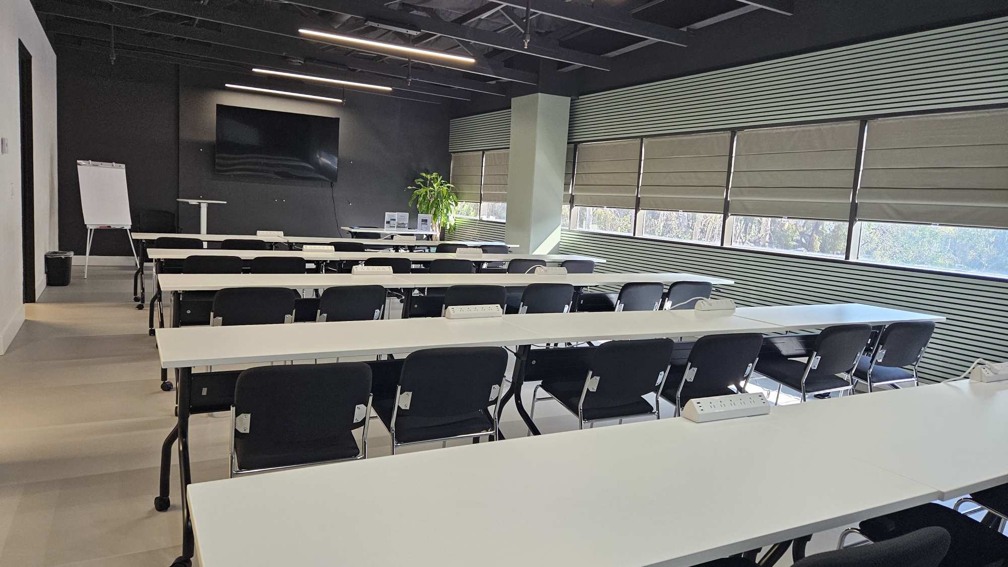 The main training room in classroom setup — rows of white tables and black chairs facing a wall-mounted screen, daylight through shuttered windows.