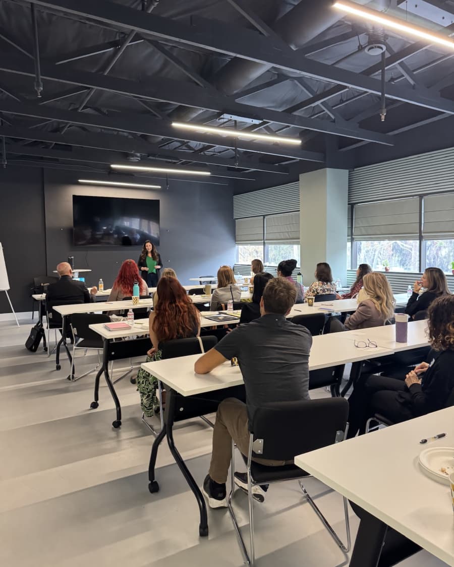 A full room of attendees seated at tables in the Encinitas training room, listening to a speaker at the front.